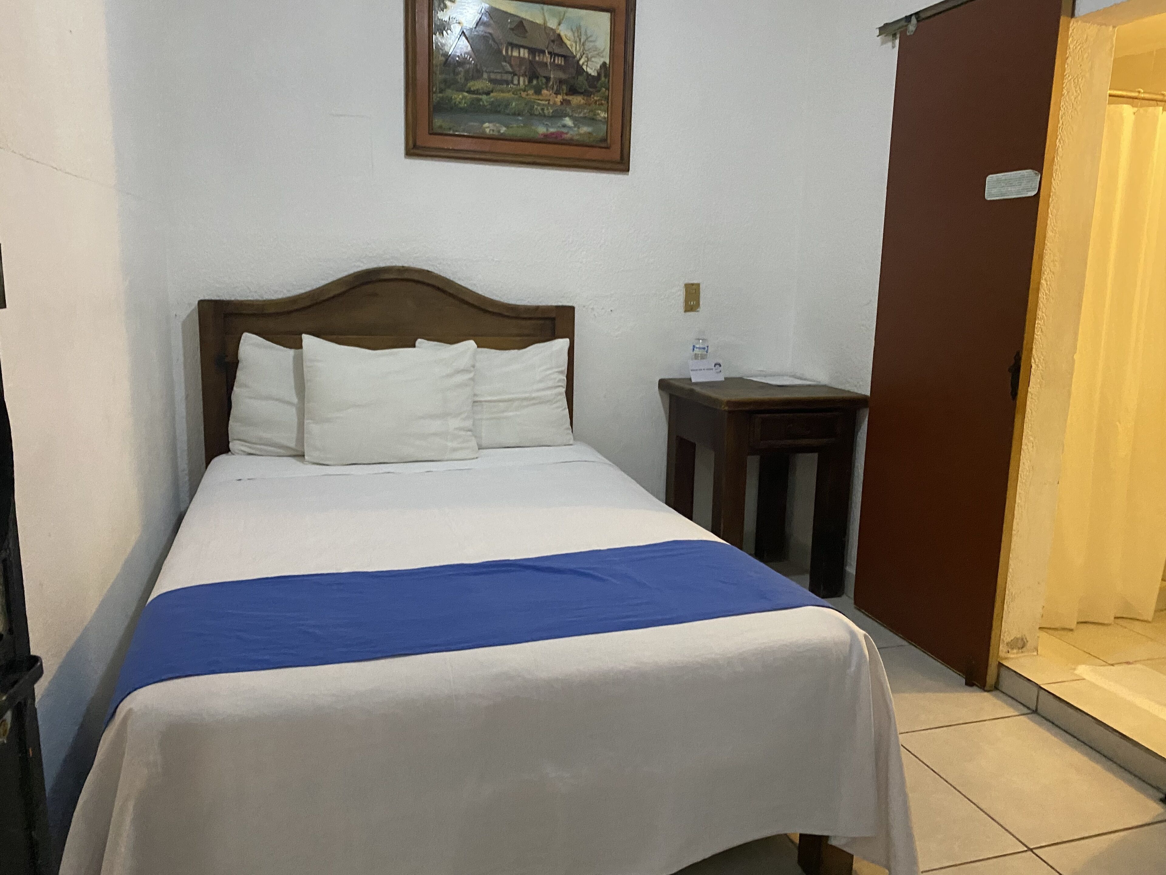 A neatly made bed with white and blue bedding, a wooden headboard, and a small bedside table with a bottle and a tissue box.