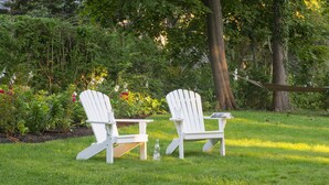 Deux chaises Adirondack blanches, accompagnées d’une petite table et d’une bouteille d’eau, sont placées dans un espace extérieur verdoyant, avec des arbres et des plantes en arrière-plan.