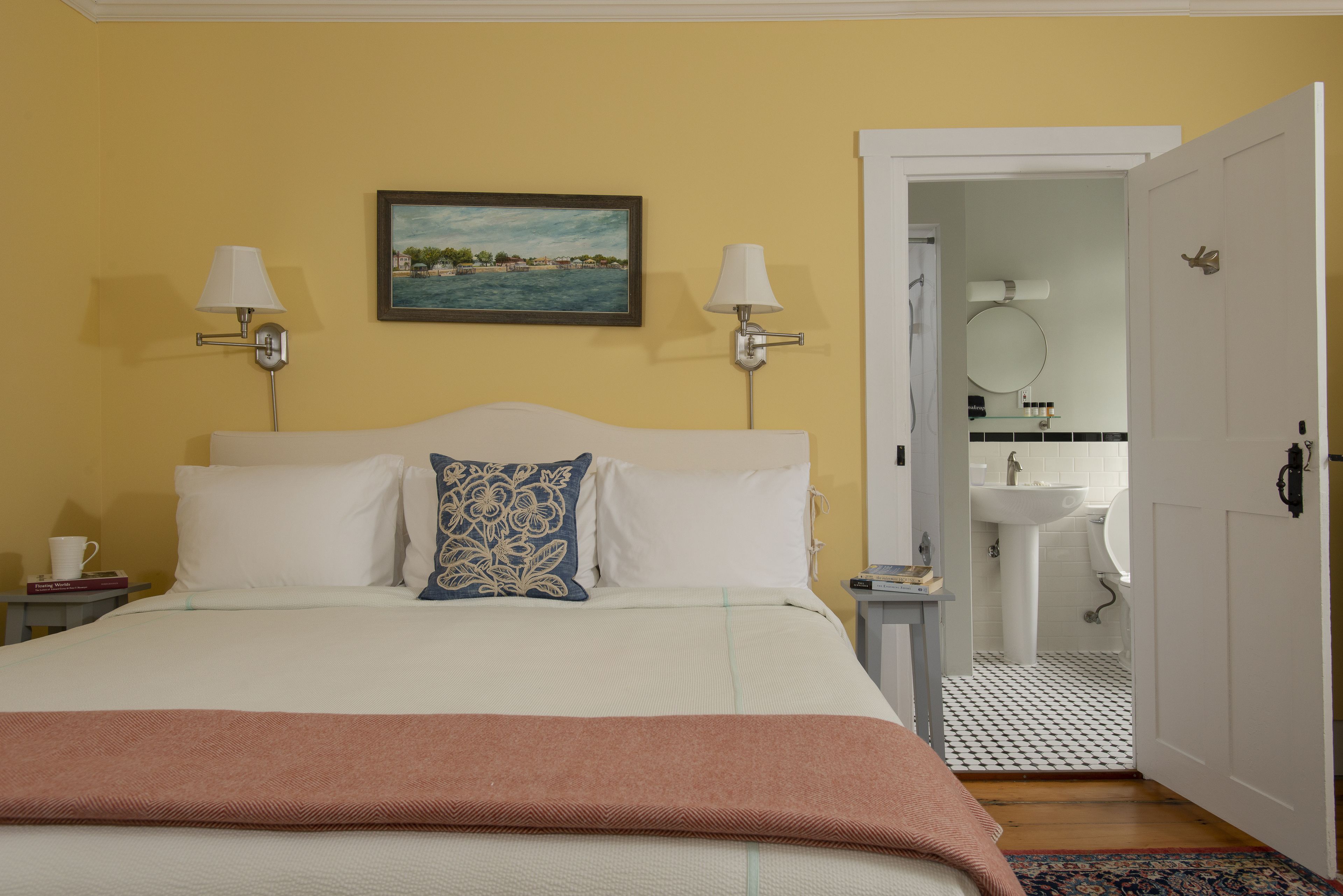 A hotel room with a bed, two bedside lamps, a painting, a nightstand, a bedside table, a white door, and a bathroom visible through an open door.