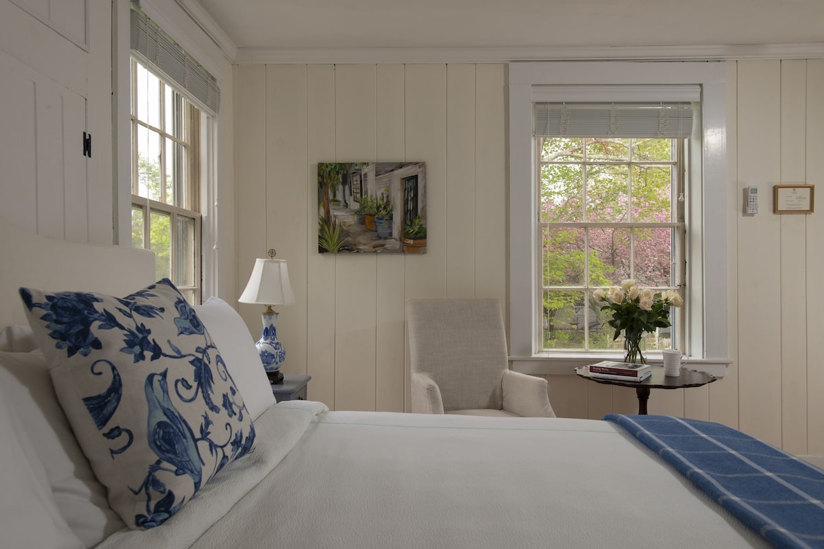 A bedroom with a large bed, a chair, a small table with a lamp, and a window with a view of trees.