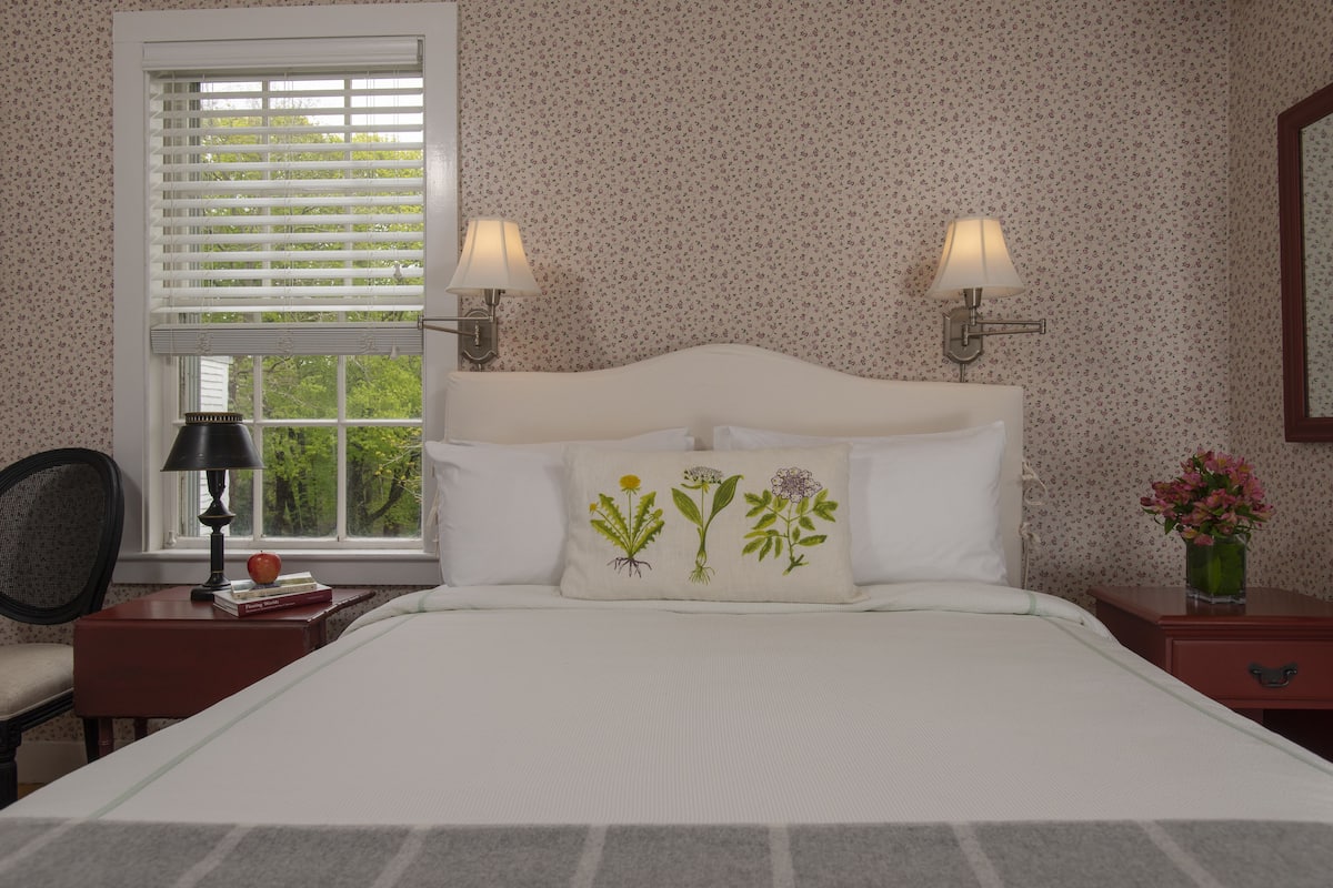 A bedroom with a bed, two bedside lamps, a window with blinds, a chair, a nightstand with a lamp, and a vase with flowers.