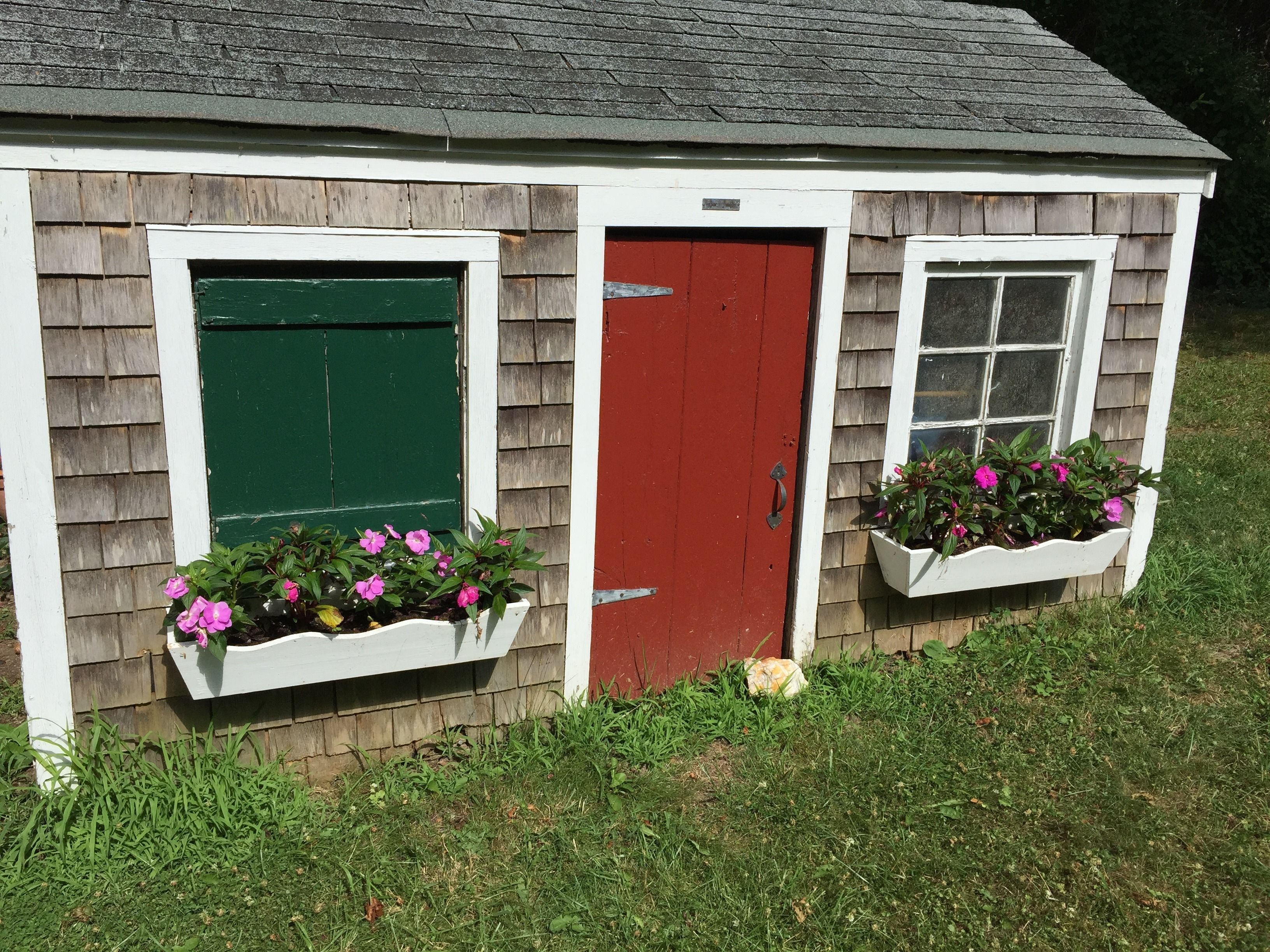 Un pequeño edificio de una sola planta con una puerta roja y dos ventanas, cada una con marco blanco y contraventanas verdes. Hay jardineras con flores rosadas a ambos lados de la puerta.