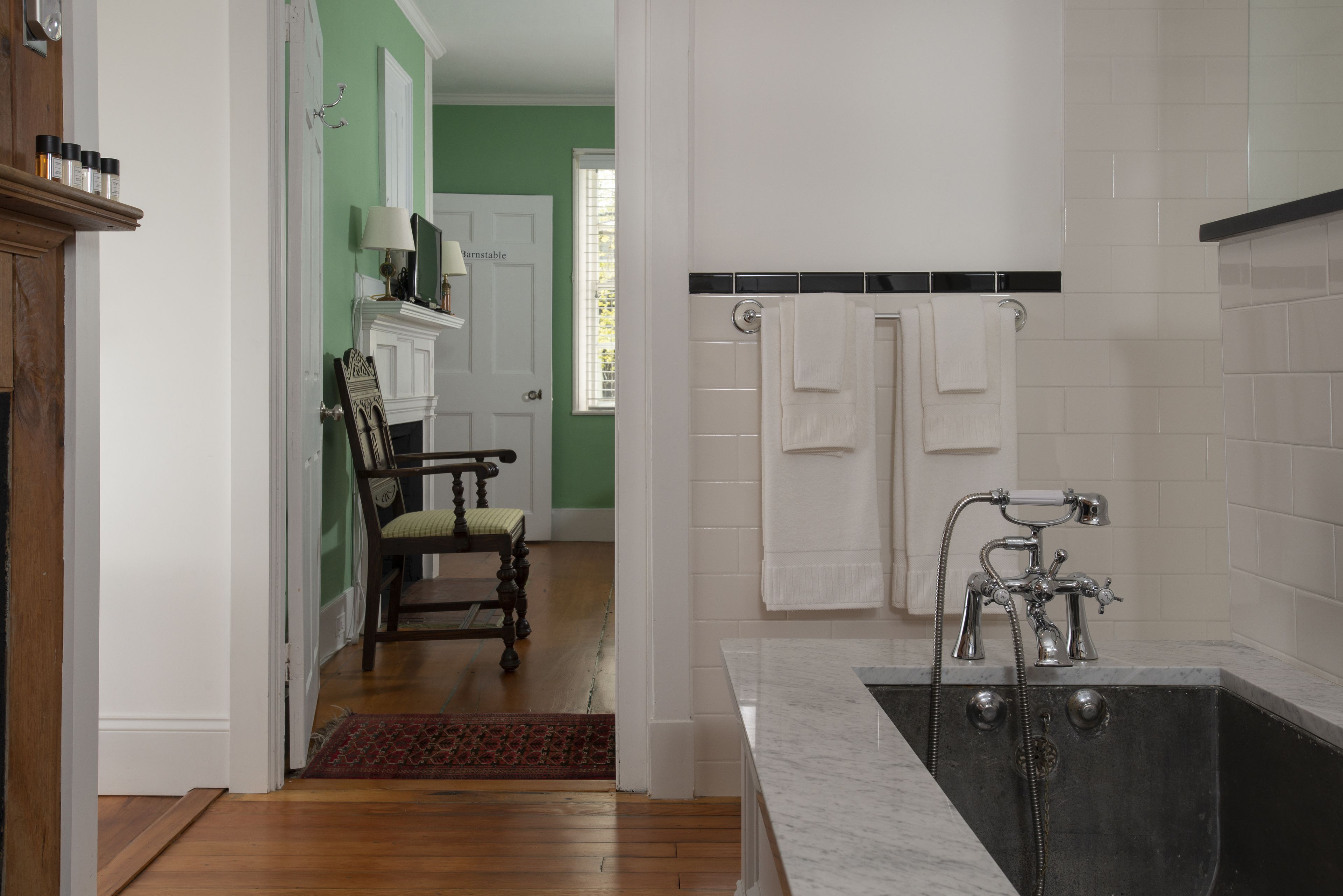 Un baño con bañera, ducha e inodoro. Se ve una puerta que da a un pasillo con una silla y una lámpara.