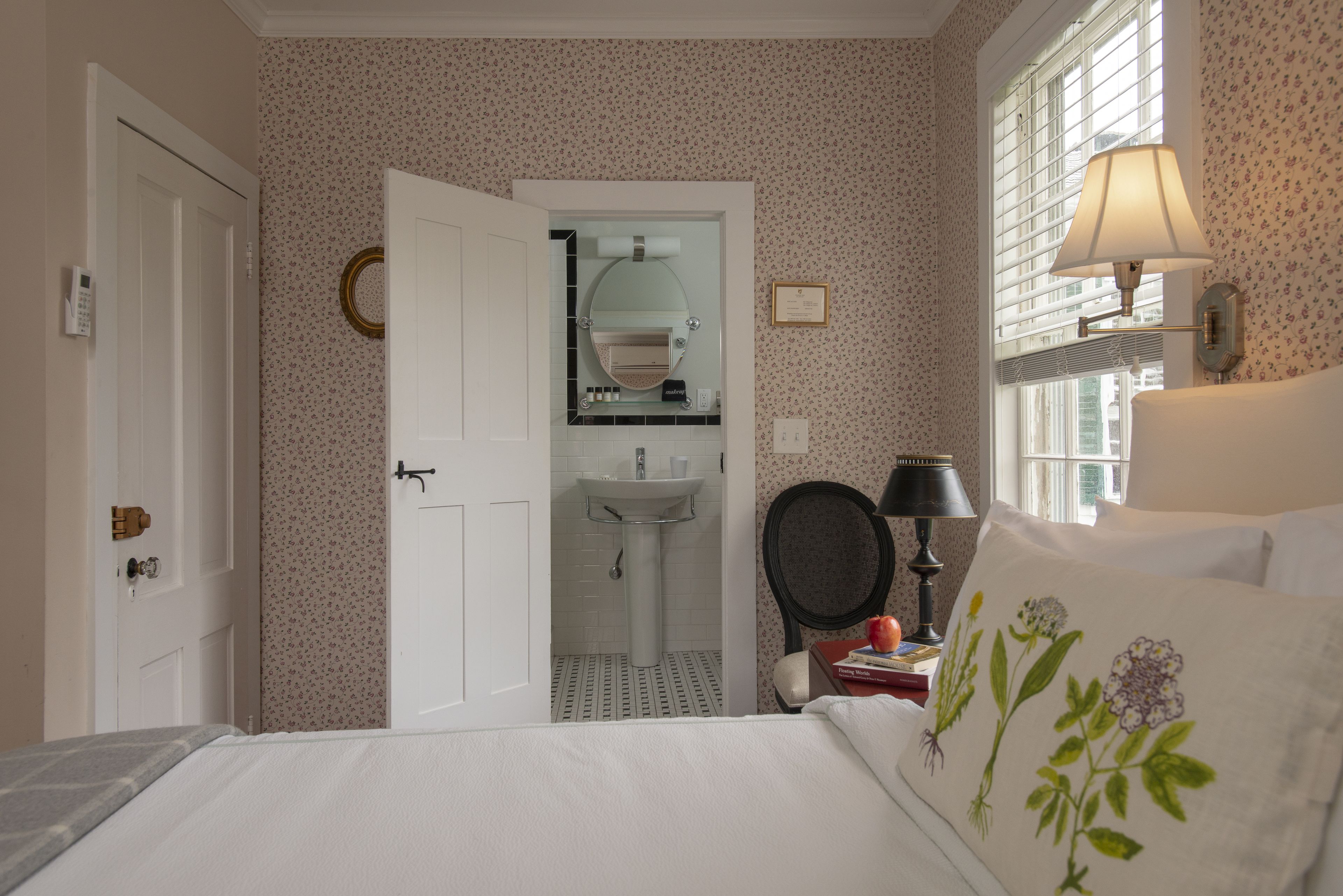 Une chambre à coucher avec un lit, une table de chevet, une lampe, une fenêtre avec des stores, et une salle de bain visible à travers une porte ouverte.