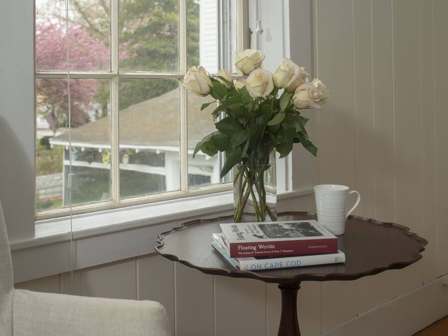 A window with a view of a house and trees, a vase with white roses, a stack of books, and a white mug on a small table.