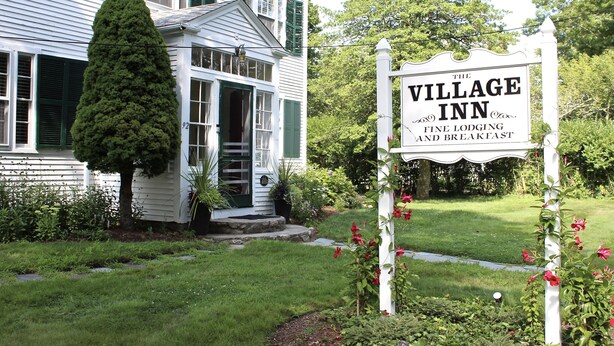 Un bâtiment blanc avec une enseigne indiquant « The Village Inn », ce qui laisse supposer qu’il s’agit d’un établissement proposant un hébergement de qualité et servant le petit-déjeuner.