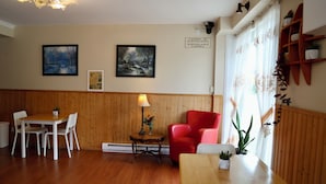 A room with wooden paneling, a dining table with chairs, a red armchair, a small table with a lamp, and two framed paintings on the wall.