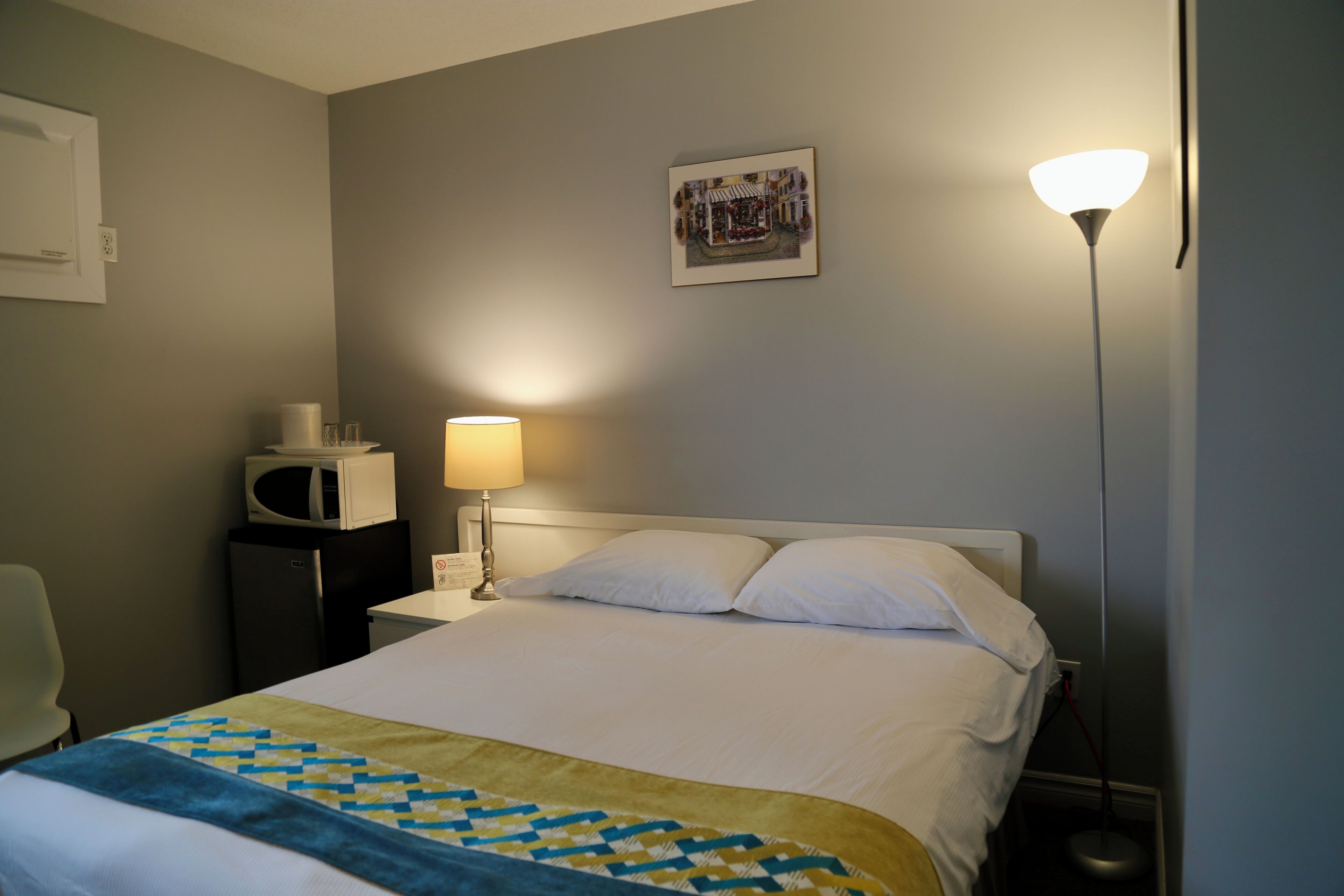 Una habitación de hotel con cama, lámpara de noche, microondas, una silla y un cuadro en la pared.