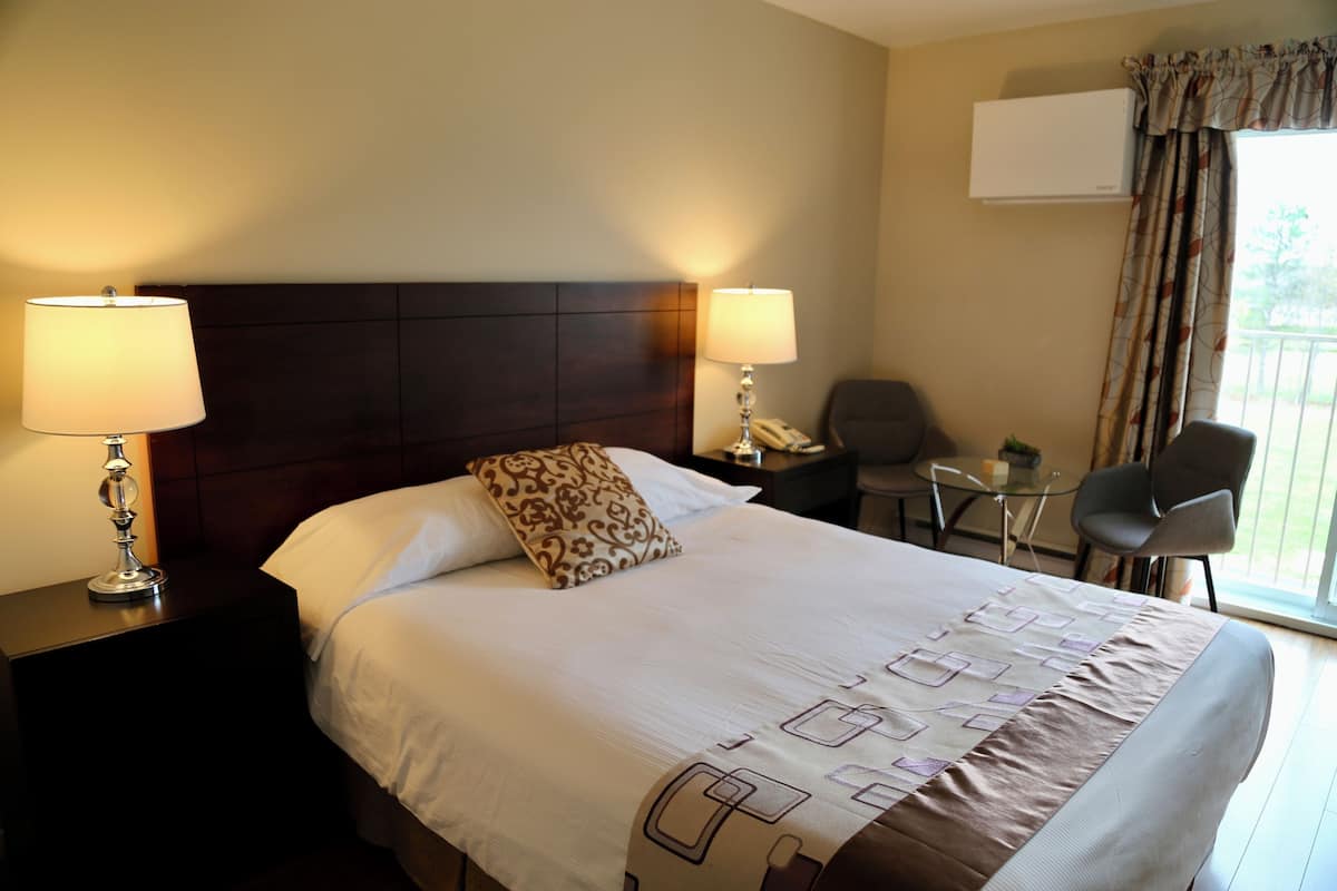A hotel room with a large bed, two bedside lamps, a small table, a chair, and an air conditioning unit.