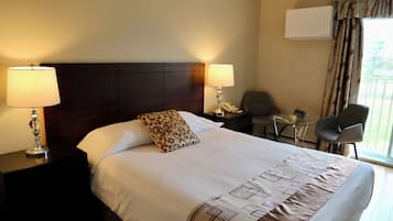 A hotel room with a large bed, two bedside lamps, a small table, a chair, and an air conditioning unit.