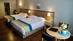 A hotel room with two beds, a desk, a chair, a small table with a vase of flowers, and an air conditioning unit on the wall.