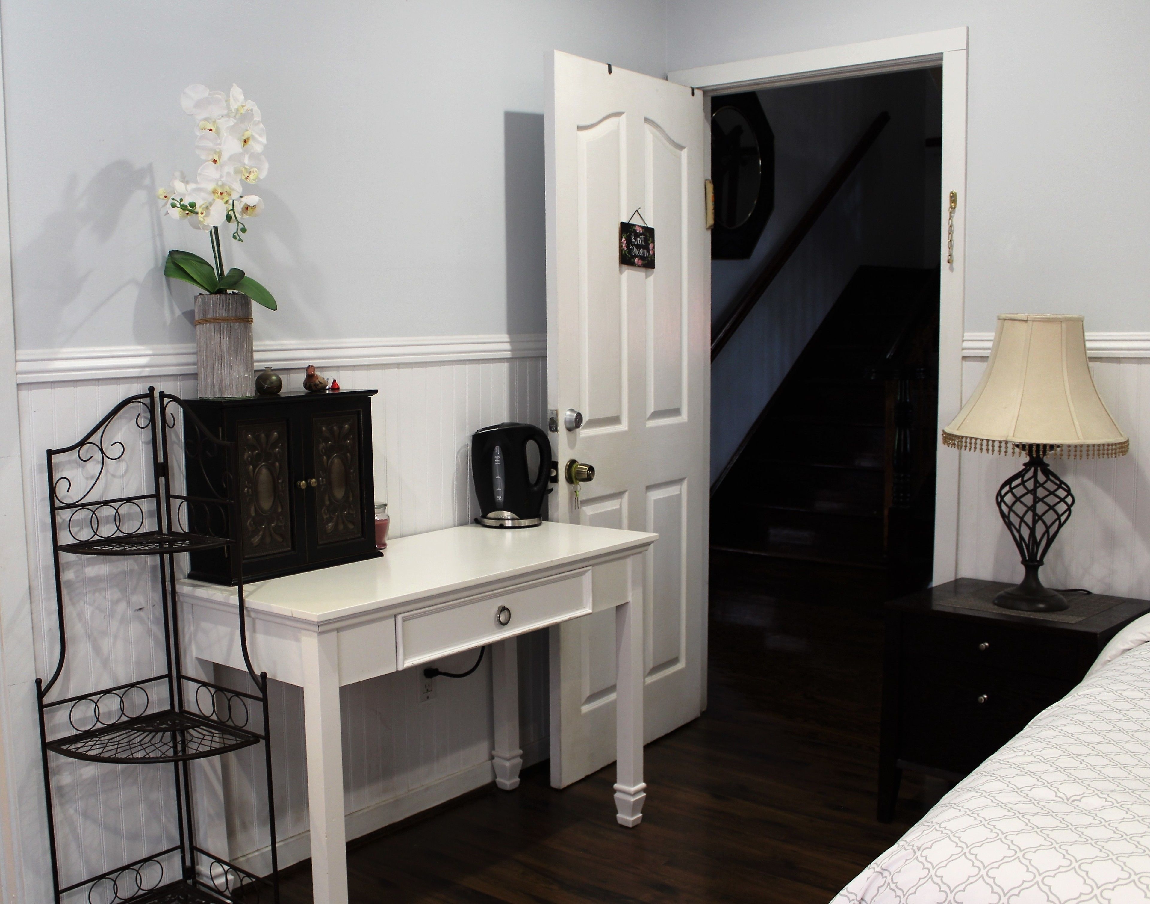 A bedroom with a white dresser, a black metal chair, a white lamp, and a staircase leading to another room.