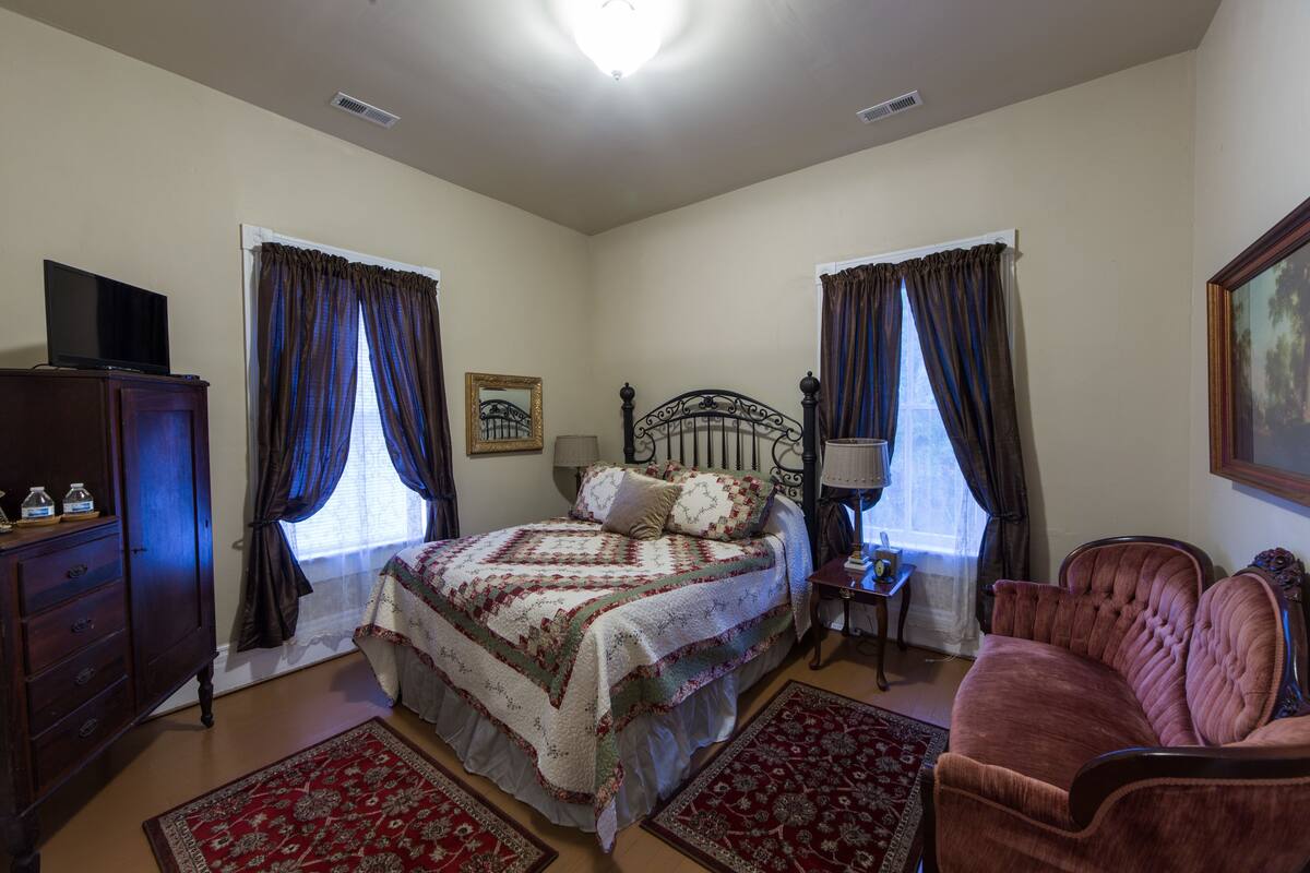 A bedroom with a bed, a dresser, a TV, a chair, and two windows with curtains.