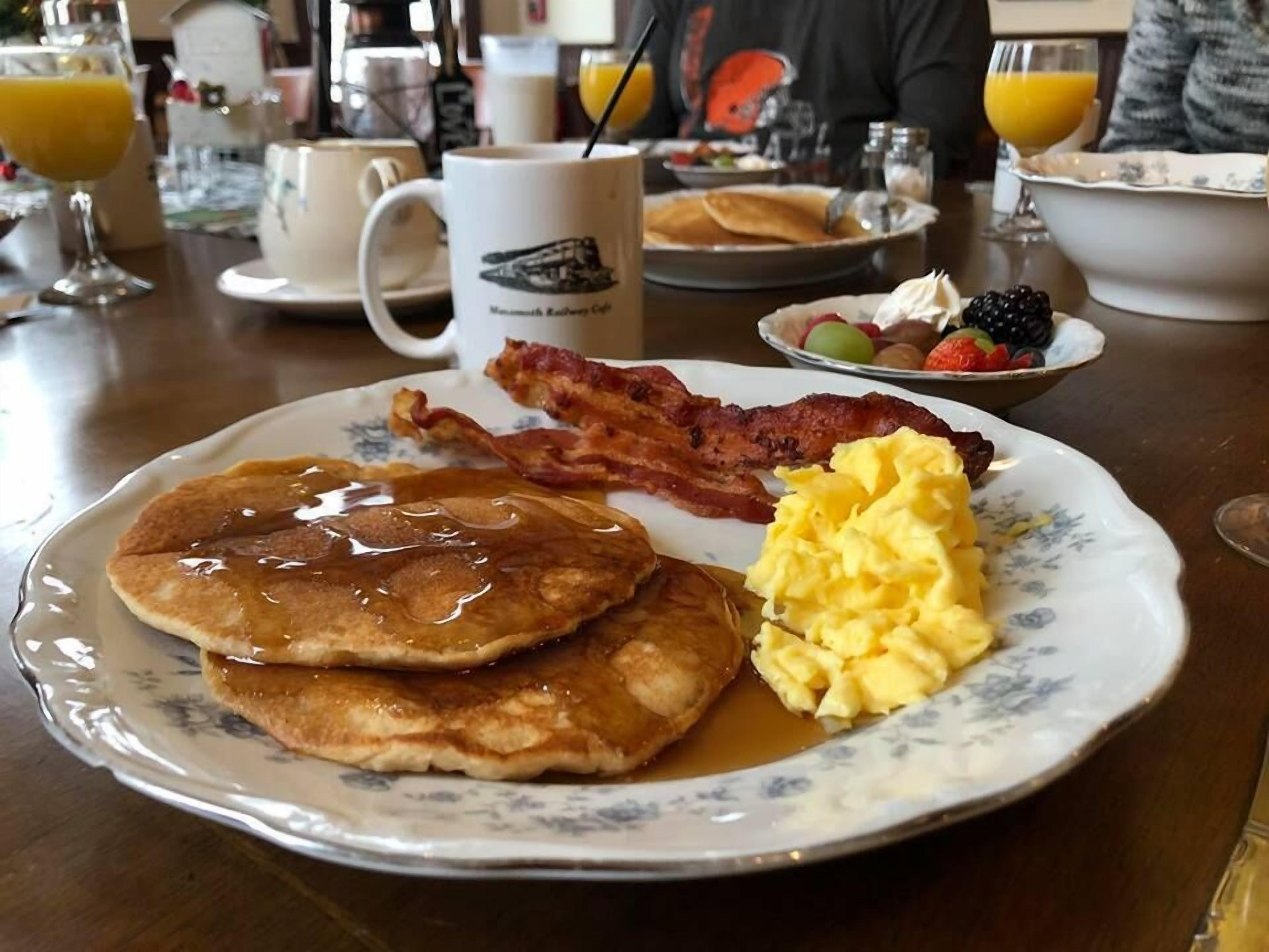 Un plato de desayuno con panqueques, tocino, huevos revueltos y fruta acompañante.