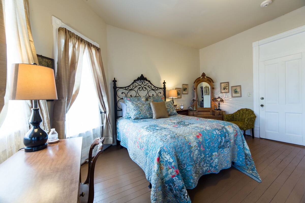 A bedroom with a large bed, a bedside table with a lamp, a chair, a dresser, and a door.