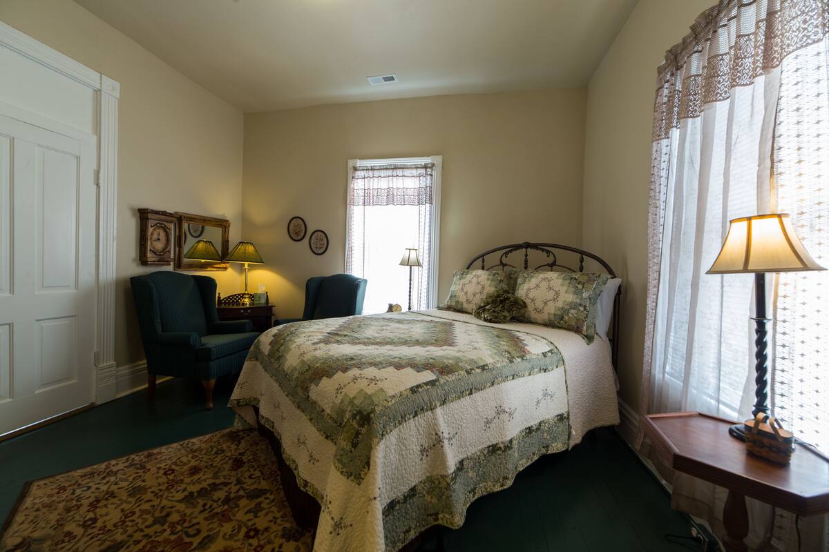 A bedroom with a bed, a chair, a nightstand, a lamp, and a window with curtains.
