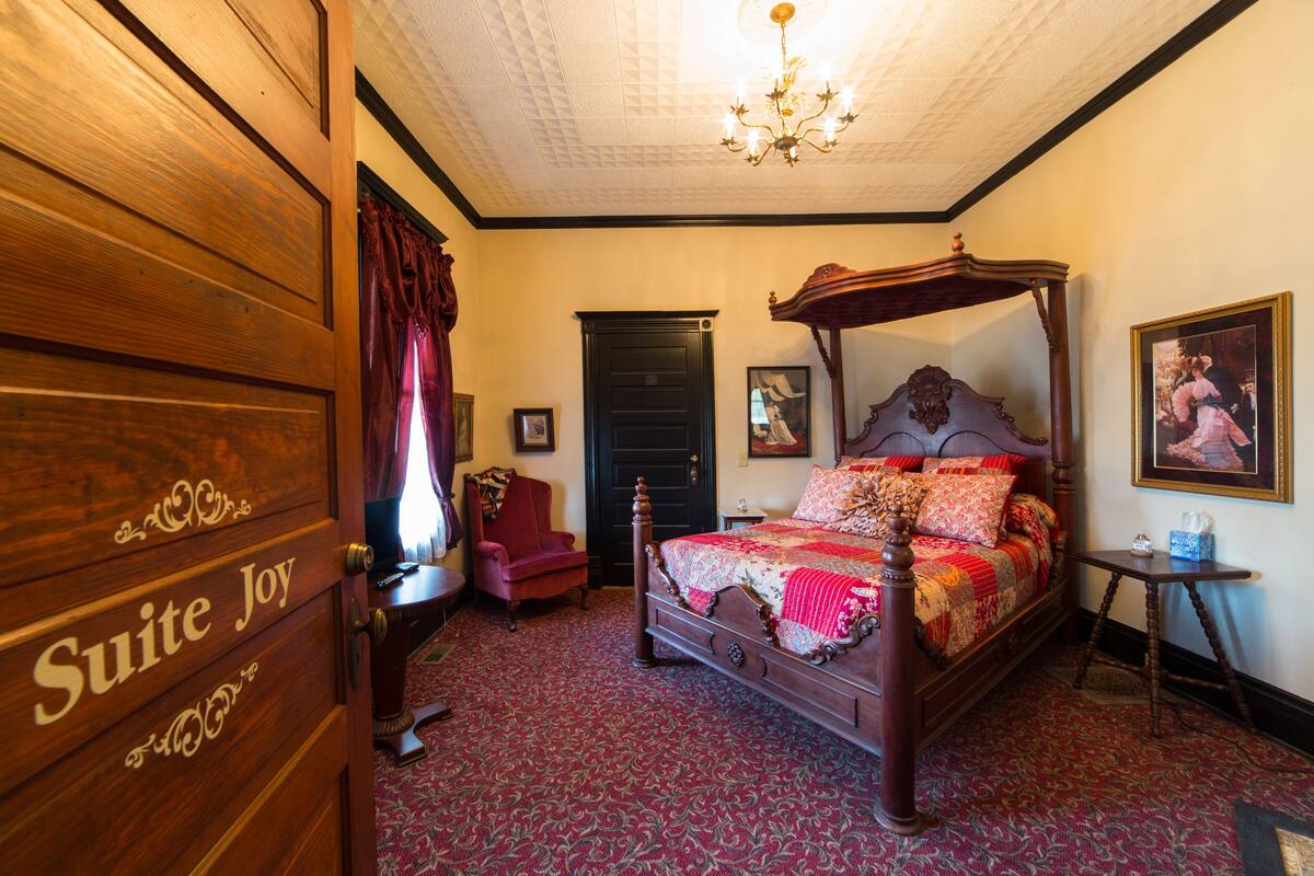 A traditional four-poster bed with a red and gold patterned bedspread, a wooden headboard, and a chandelier above.