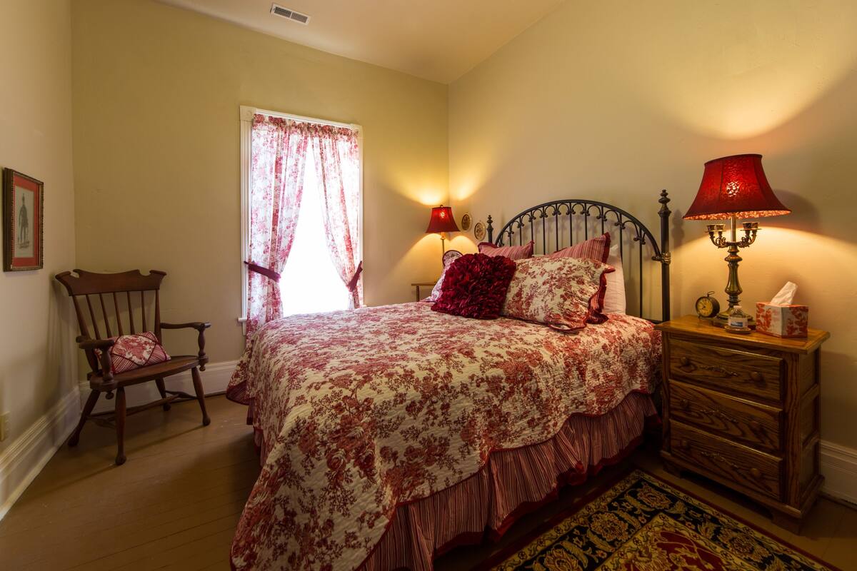 A bedroom with a bed, a nightstand, a lamp, a chair, and a window with curtains.