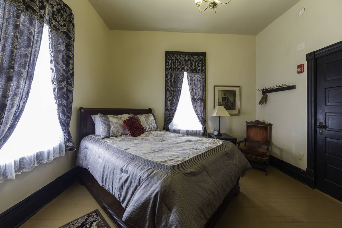 A bedroom with a bed, two windows with curtains, a nightstand, a lamp, a coat rack, and a door.