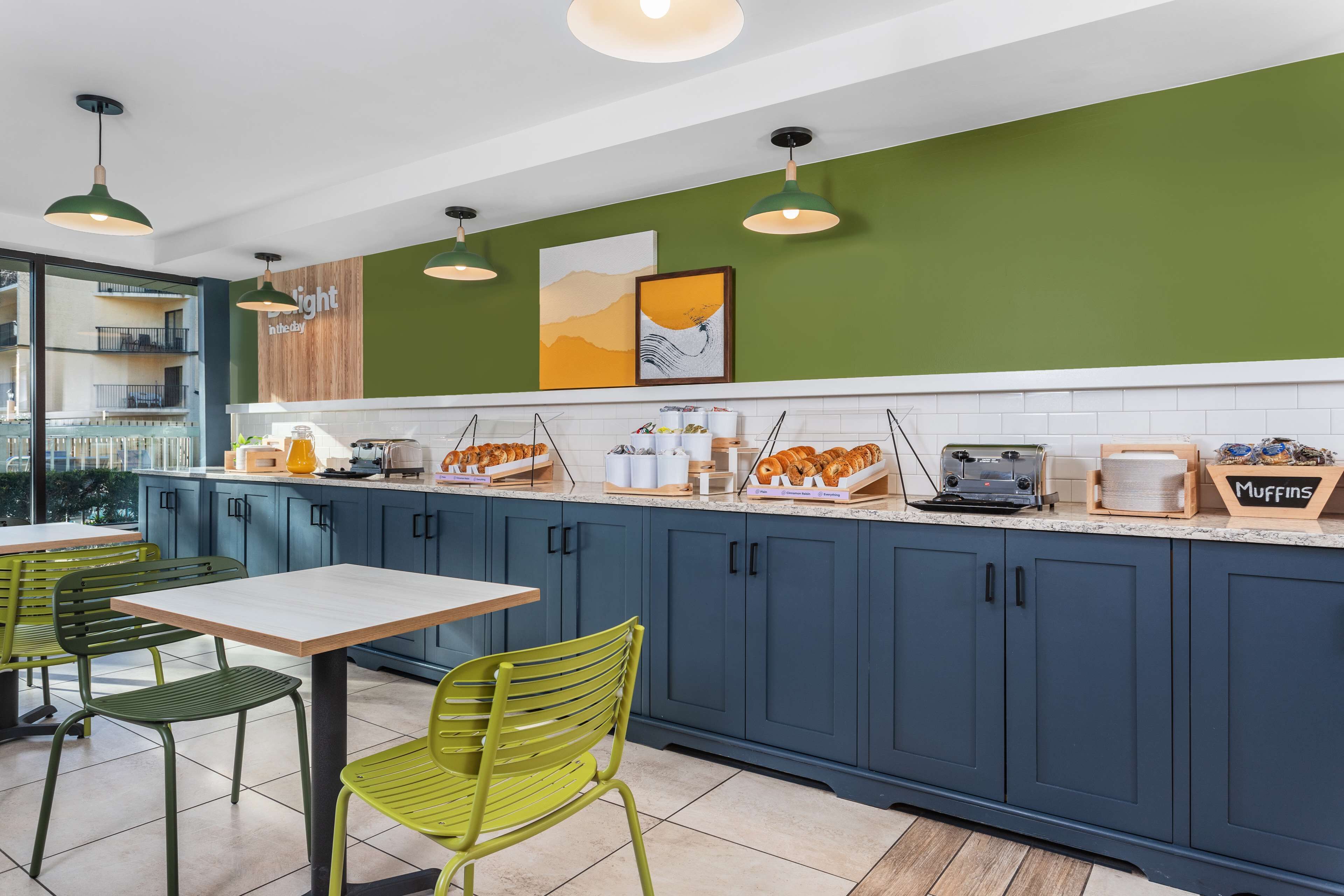 Una cocina moderna con armarios azules, encimera blanca y sillas verdes. Se observan varios alimentos sobre la encimera, como pasteles y una cafetera.