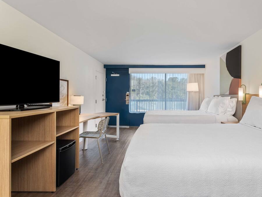 A hotel room with two beds, a desk, a television, and a window with a view of trees.