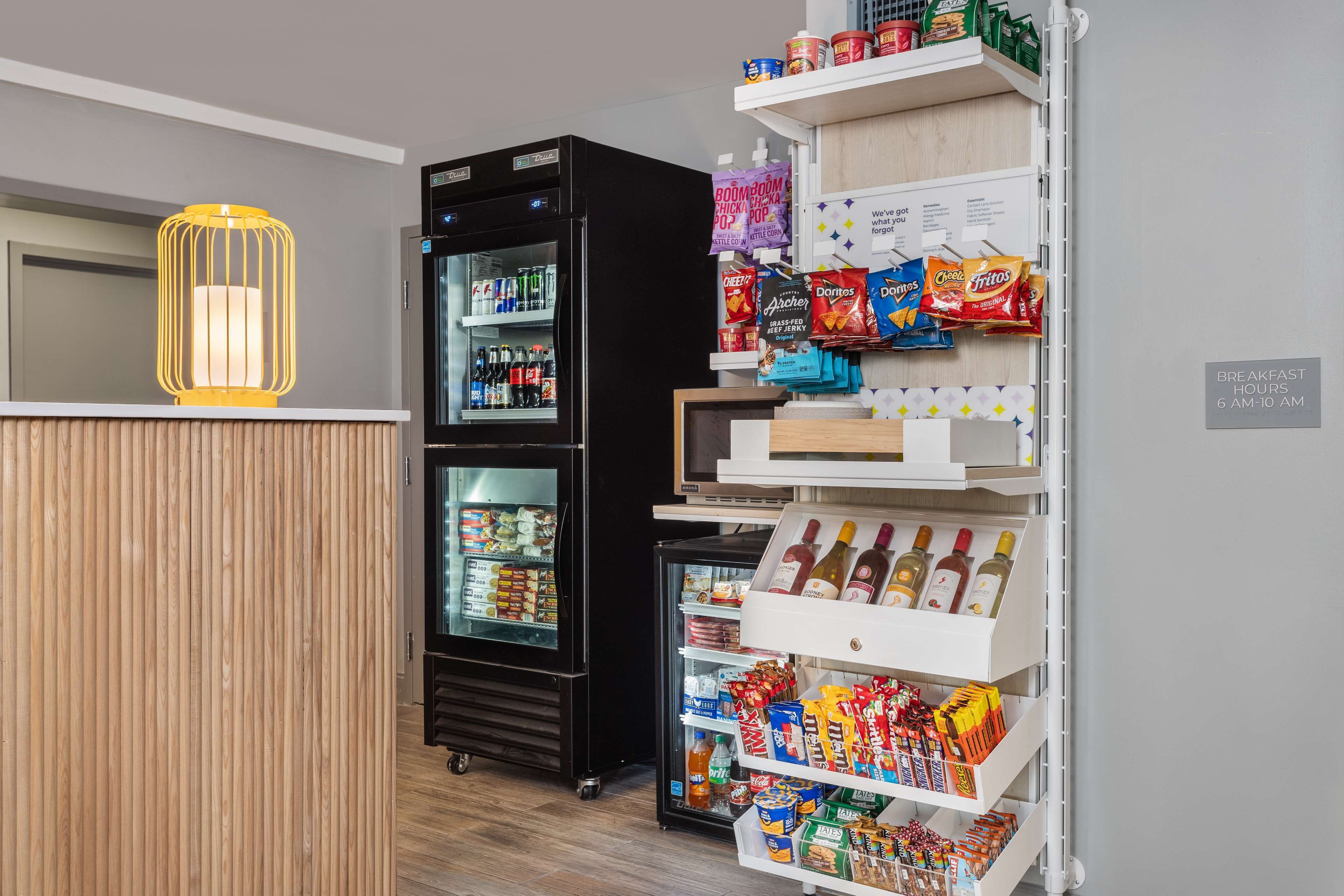 A hotel lobby with a wooden reception desk, a black refrigerator stocked with beverages, snack shelves, and a wine cooler.