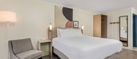 A modern hotel room with a large bed, a chair, a bedside table, and a wall-mounted lamp.