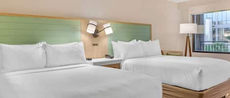 Habitación de hotel con dos camas, cabecera verde, lámpara de noche y ventana con vista a un edificio.