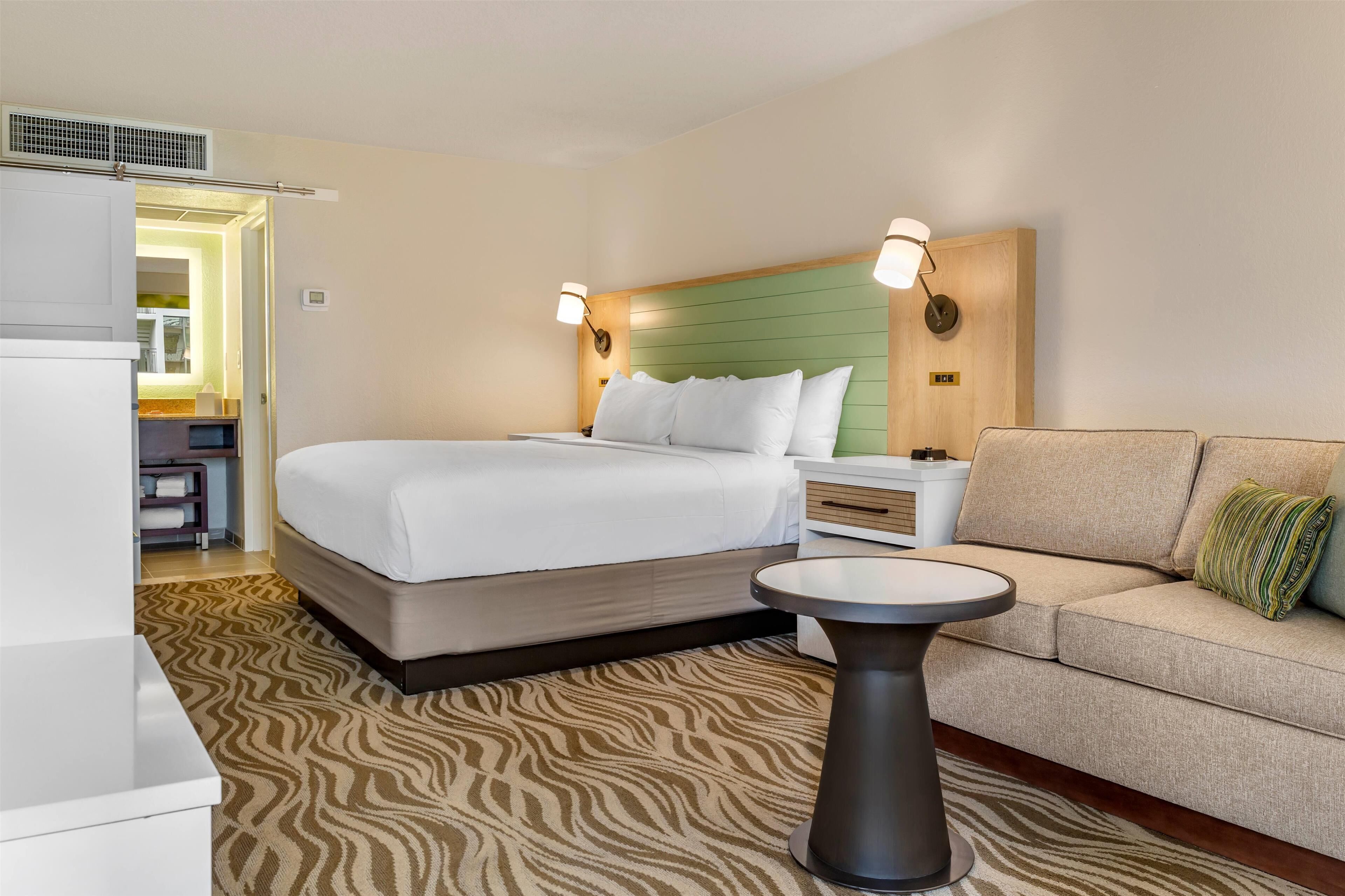 Quarto de hotel com cama, mesa de cabeceira, sofá, uma pequena mesa e tapete estampado.