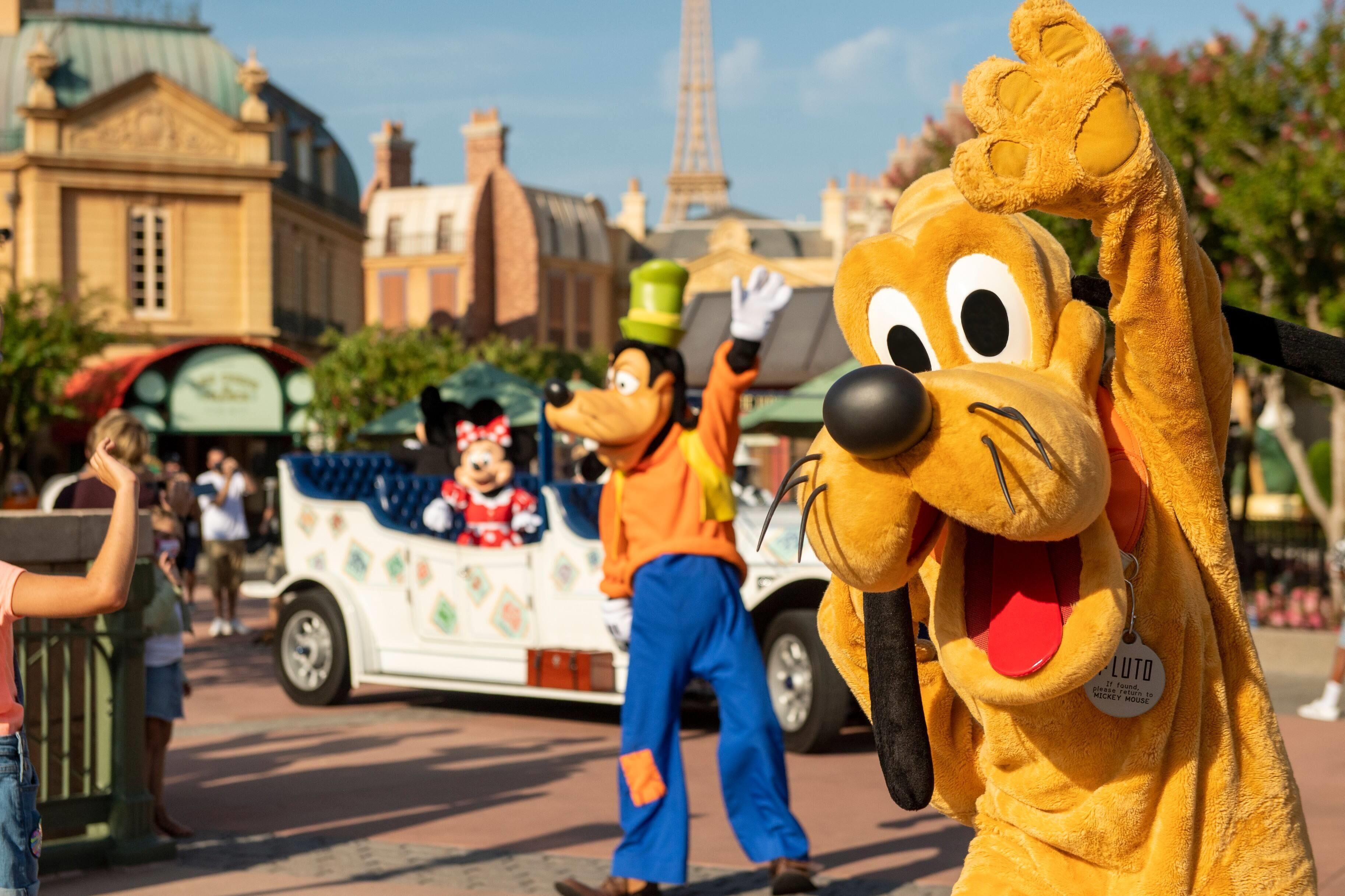 A character in a yellow costume with a green hat and a red bow tie, standing in front of a white vehicle with colorful designs and a character in a blue outfit.