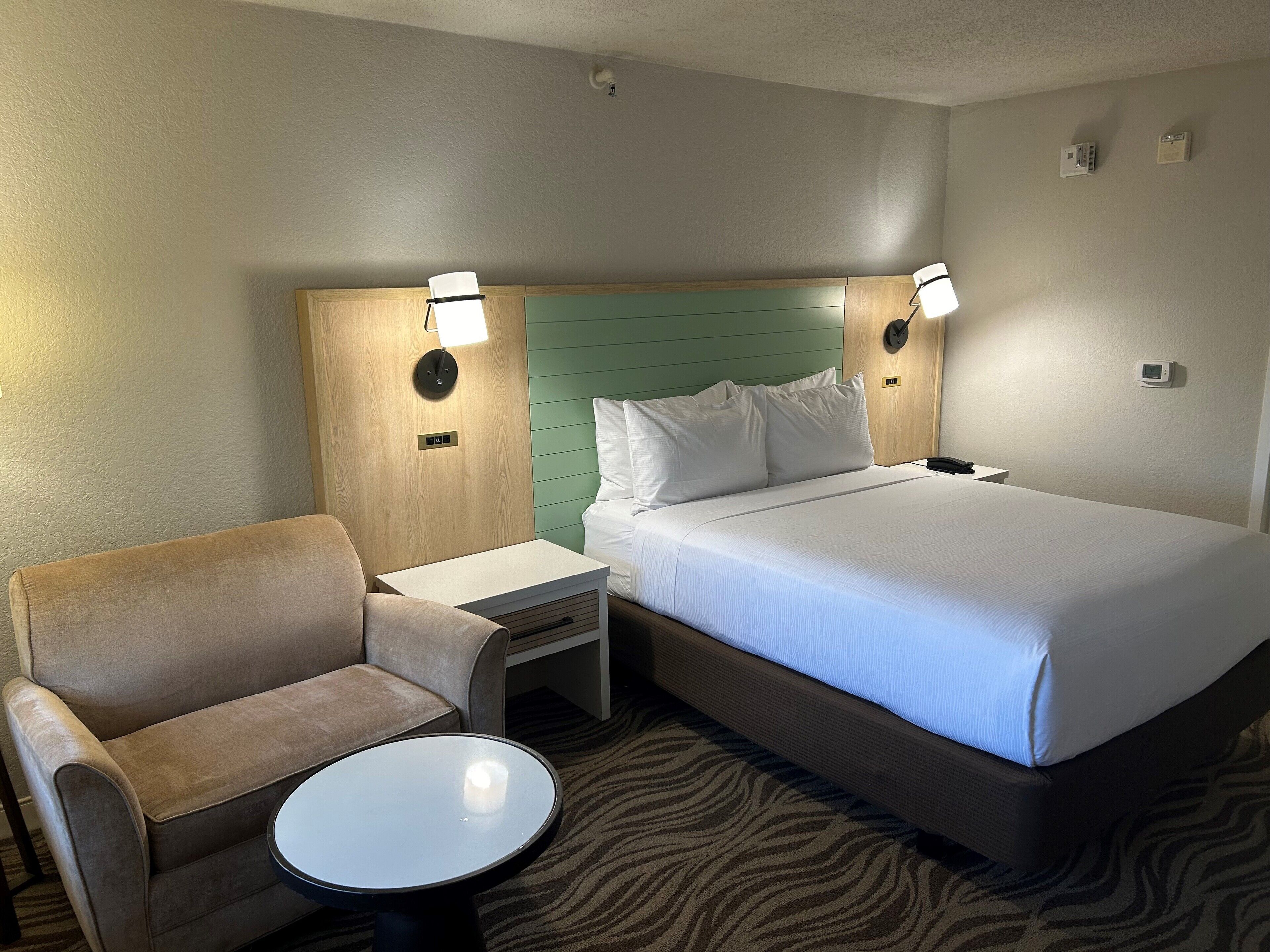 A hotel room with a bed, a chair, a small table, and two wall-mounted lamps.