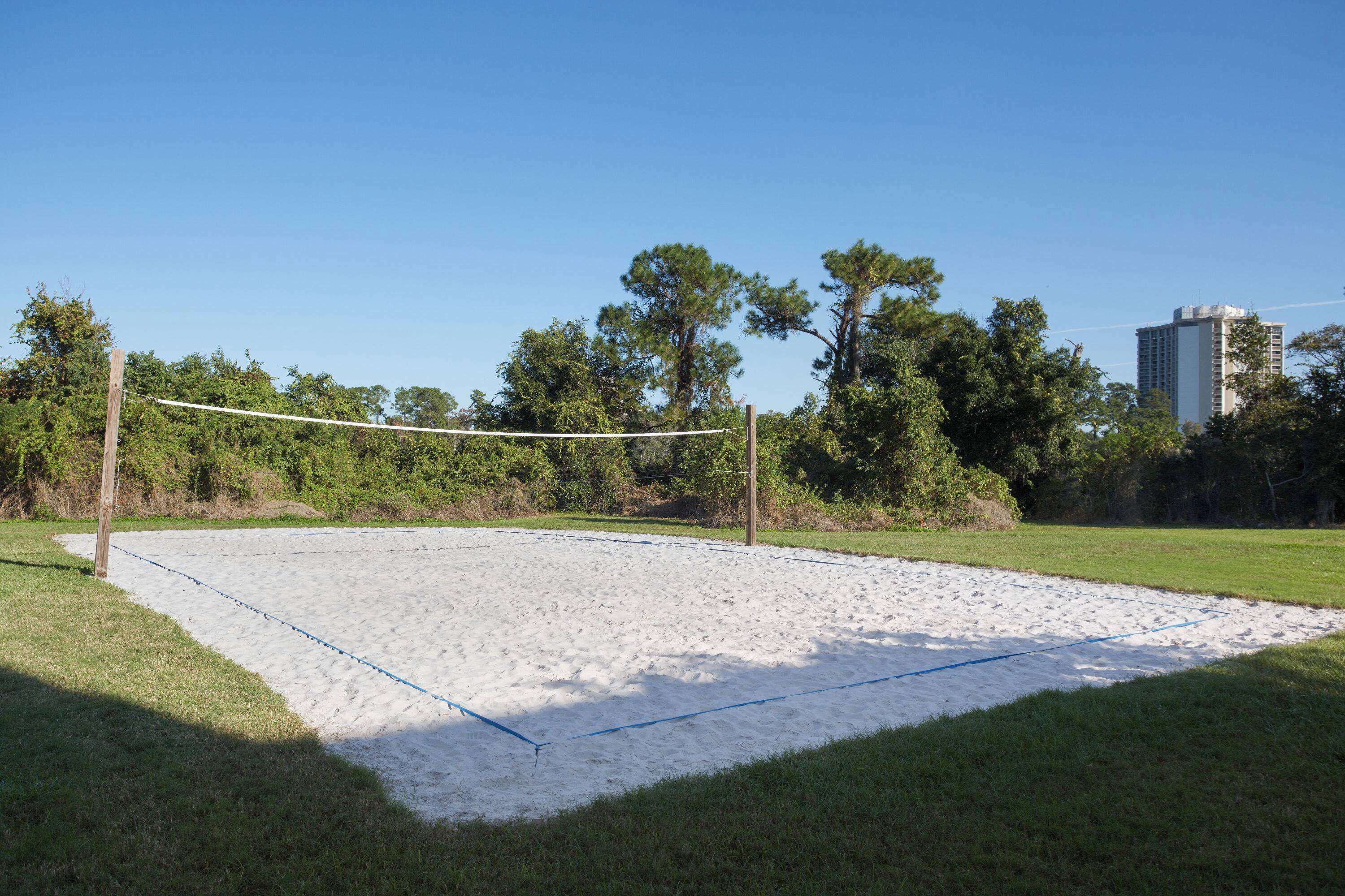 Una cancha de voleibol de arena con una red, rodeada de césped y árboles, con un edificio alto al fondo.