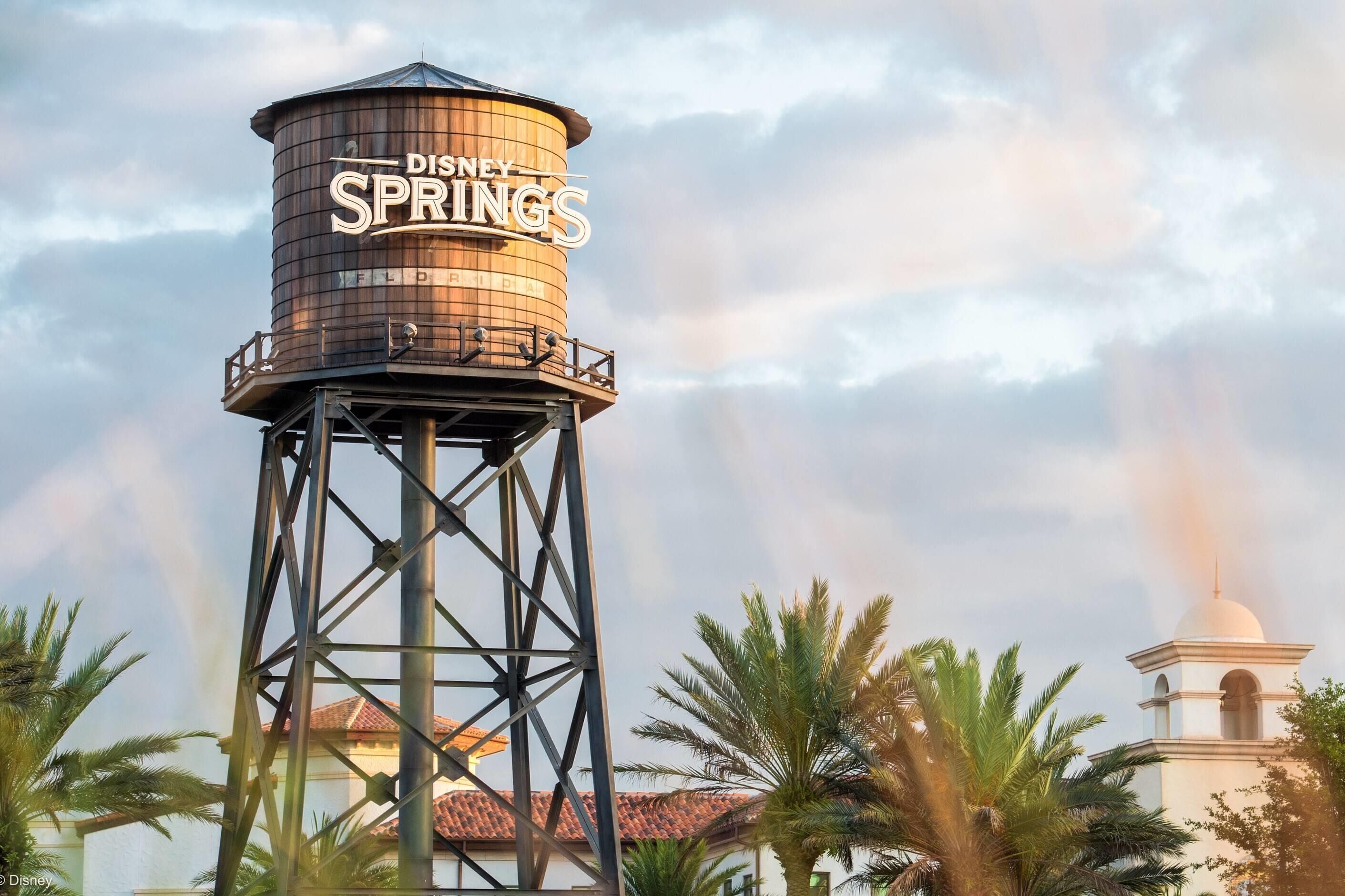 A water tower with the Disney Springs logo, surrounded by palm trees and a building with a red-tiled roof.