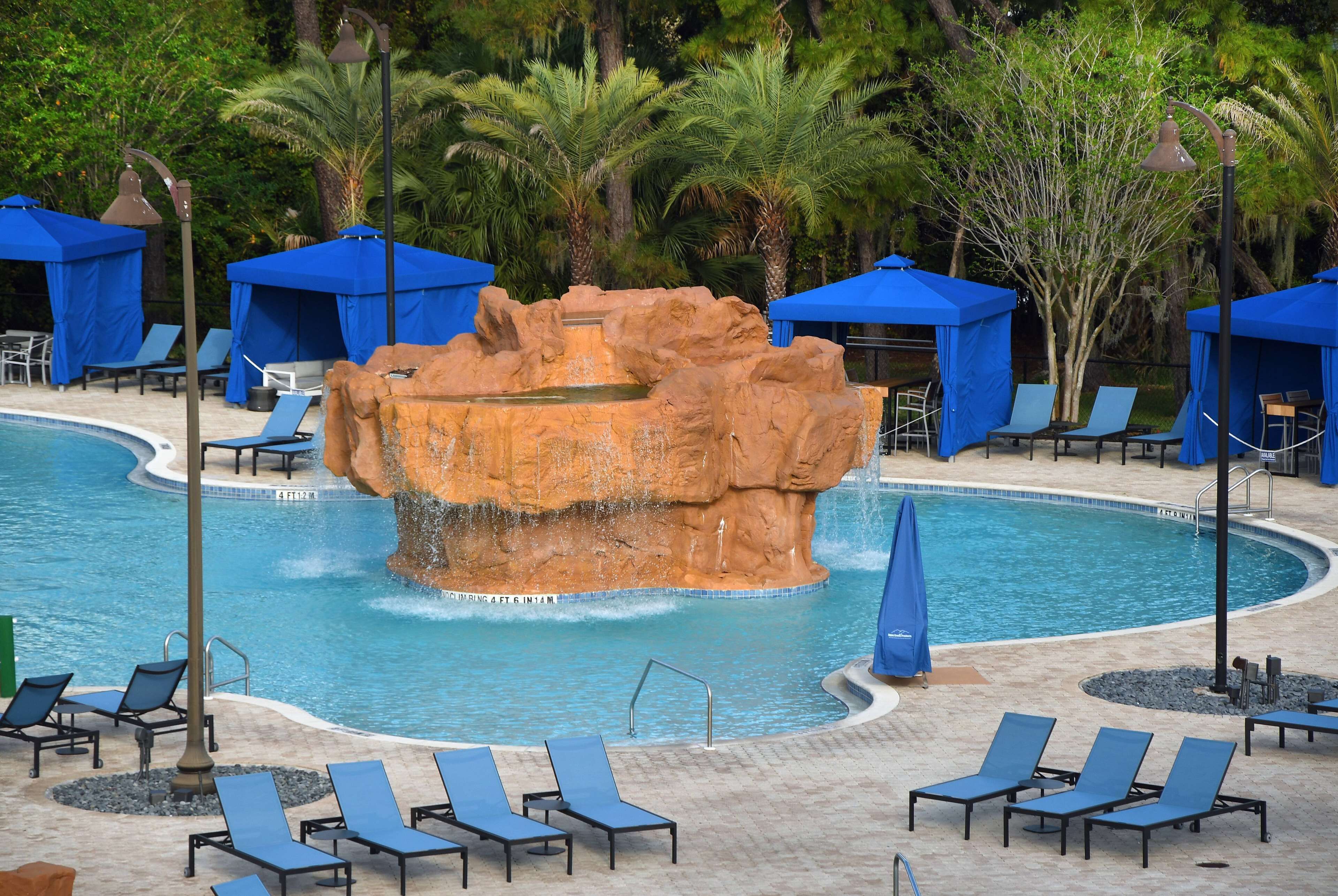 Zona de piscina con sillas reclinables azules, una formación rocosa en el centro y sombrillas azules.