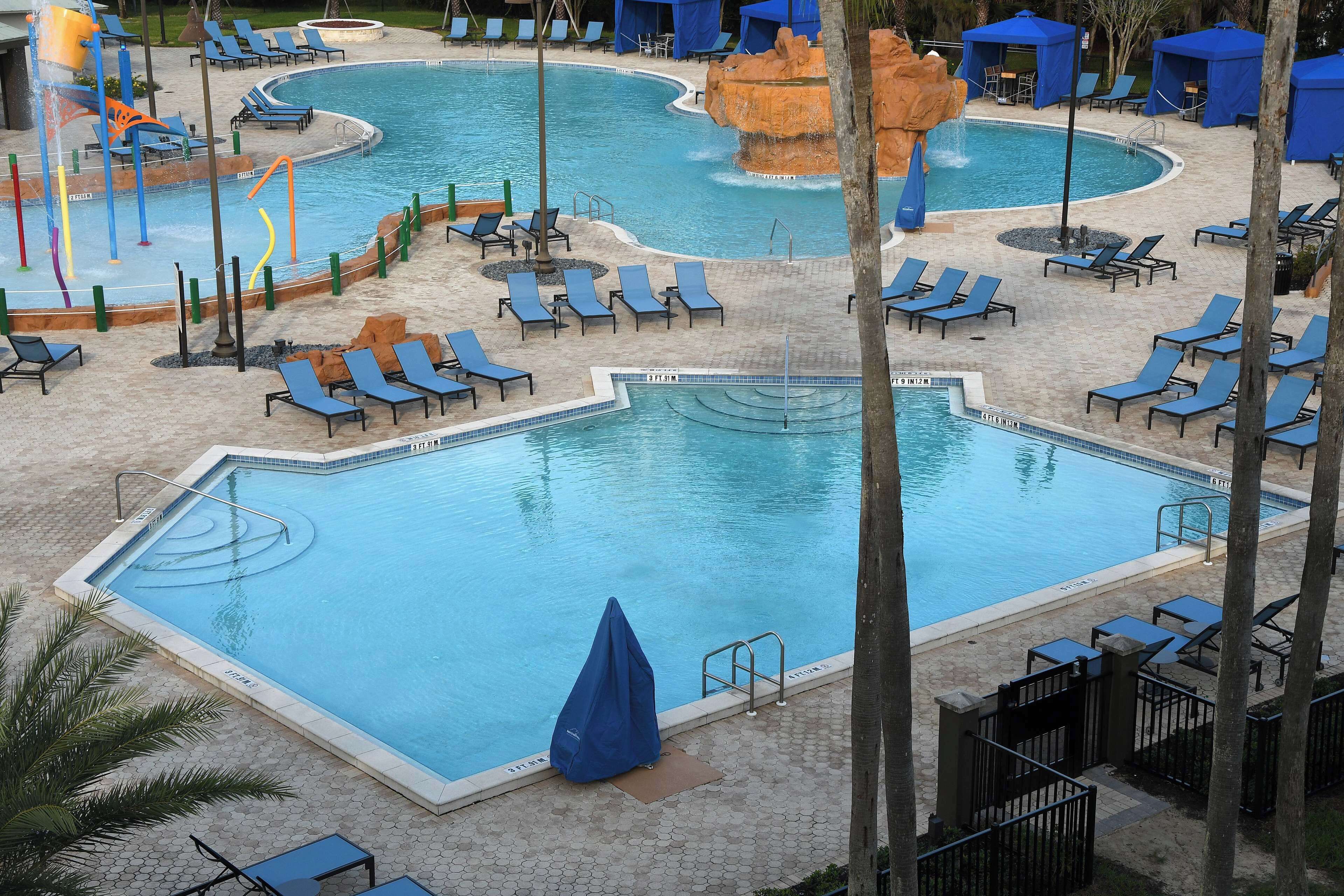 A large pool with multiple sections, including a central area and a smaller pool with a waterfall feature. There are numerous lounge chairs and umbrellas around the pool area.