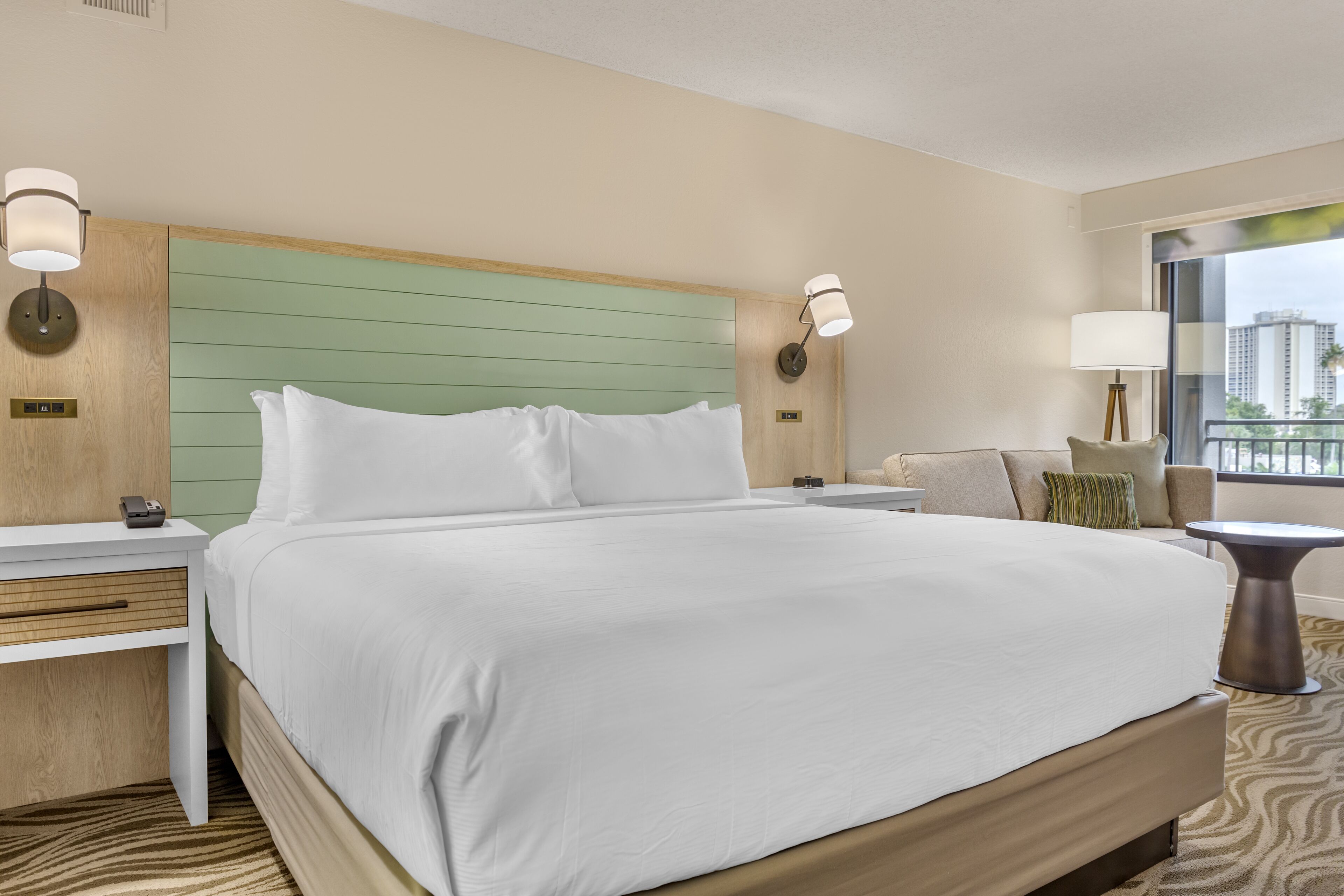 A hotel room with a large bed, wooden headboard, bedside tables, and a view of buildings through the window.