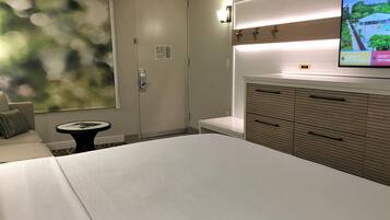 Quarto de hotel com cama, uma mesinha, televisão fixada na parede e porta com puxador.