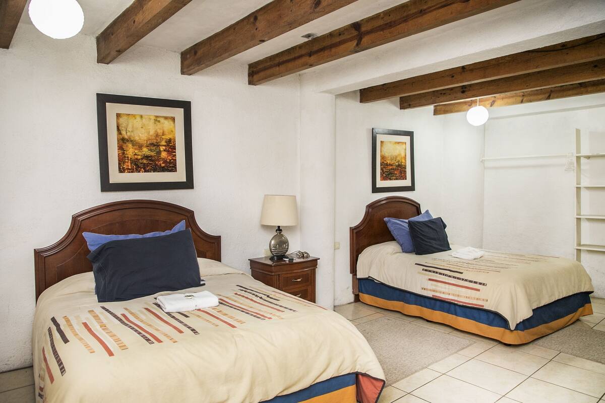 Habitación de hotel con dos camas, cabeceros de madera, una mesita de noche con lámpara y cuadros en las paredes.