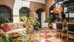 A hotel lobby with a checkered floor, a sofa with a red pillow, a glass coffee table, and several potted plants.