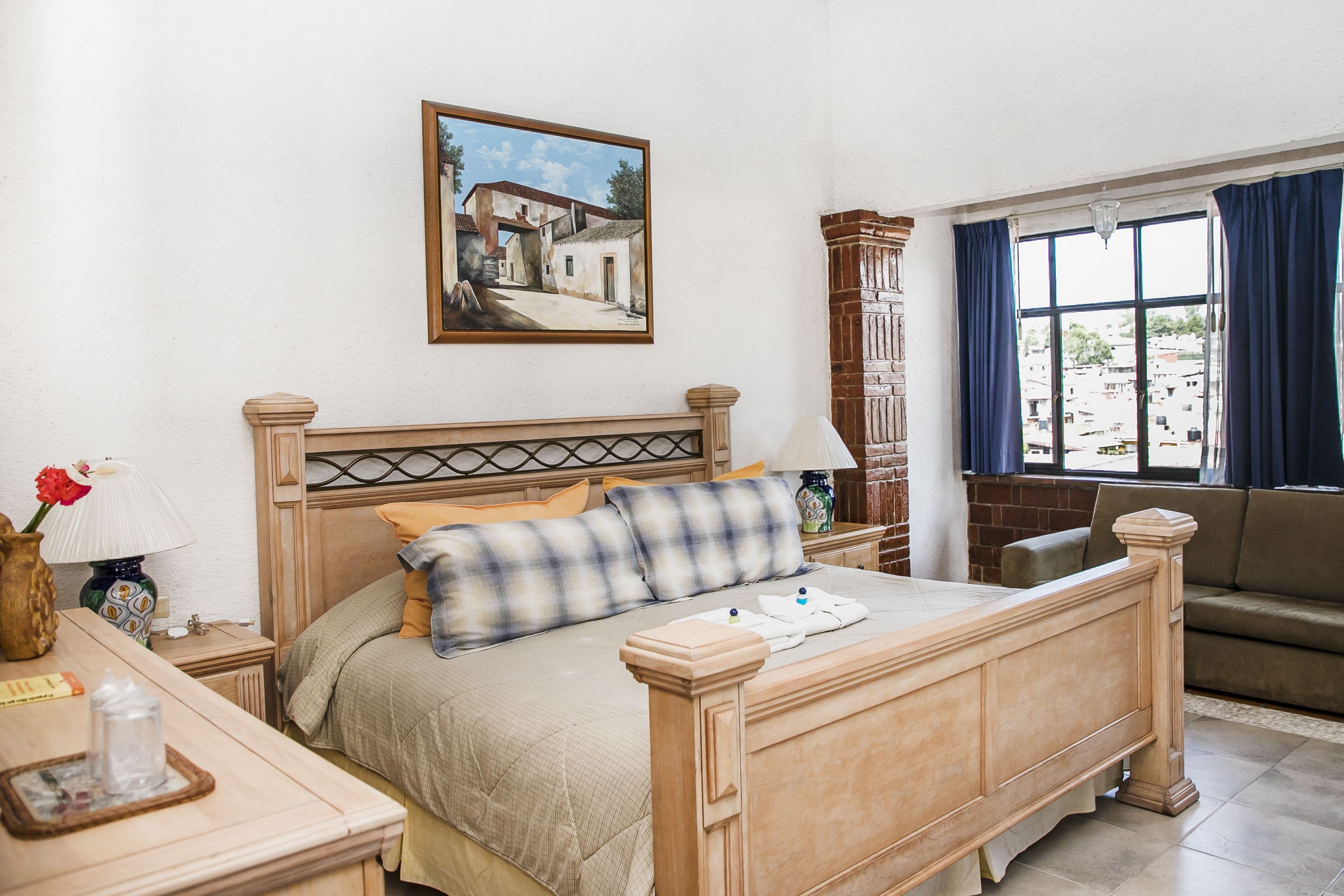 A neatly arranged bedroom with a wooden bed, a painting on the wall, and a view of the city through a window.