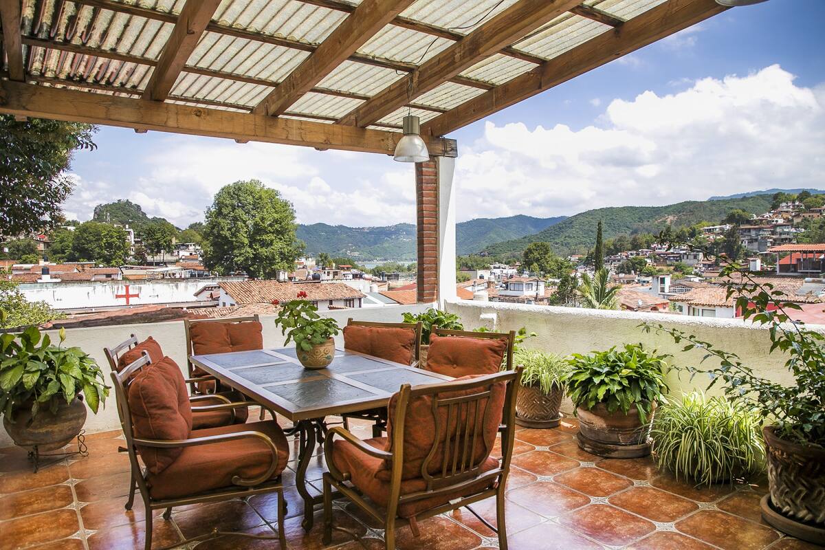 Un porche cubierto con una mesa y sillas, rodeado de plantas en macetas, con vistas a un pueblo de edificios con tejados rojos y colinas verdes.