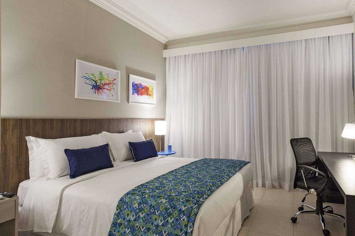 A hotel room with a bed, a desk, a chair, a TV, and artwork on the wall.