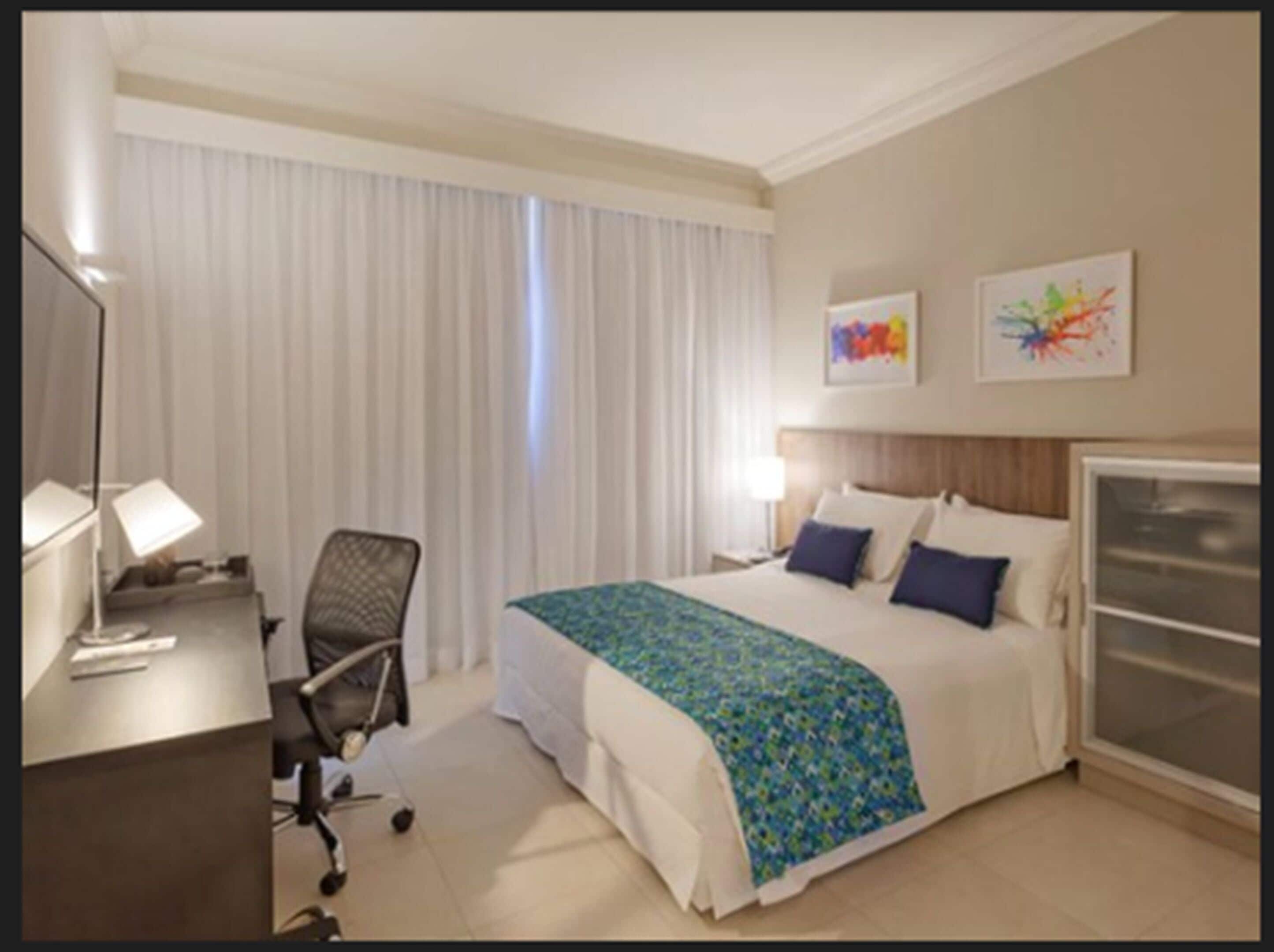 A hotel room with a bed, a desk with a lamp, a chair, and a large window with curtains.