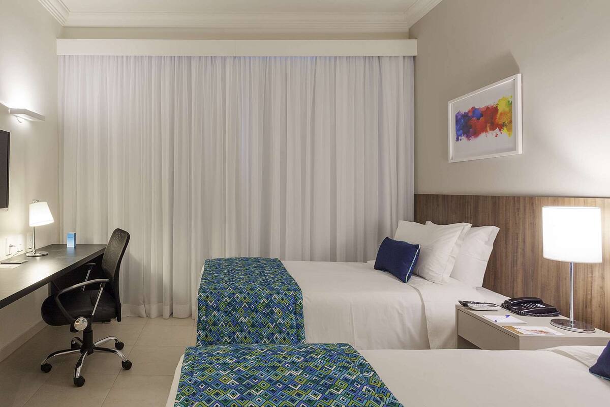 A hotel room with a desk, two beds, a TV, and a painting on the wall.