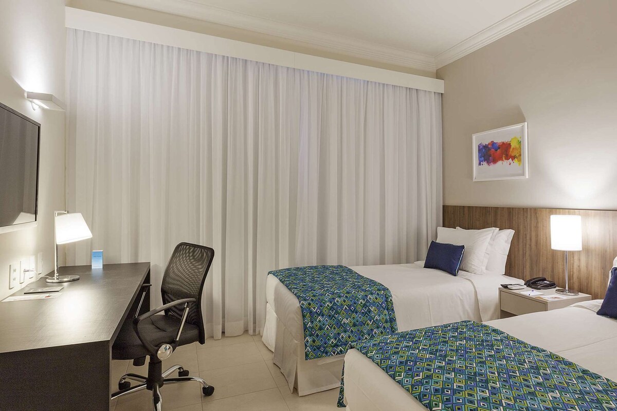 Habitación de hotel con dos camas, un escritorio, una silla y una mesita de noche con lámpara.