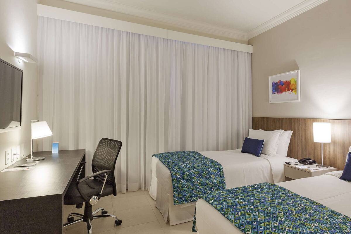 A hotel room with two beds, a desk, a chair, and a nightstand with a lamp.