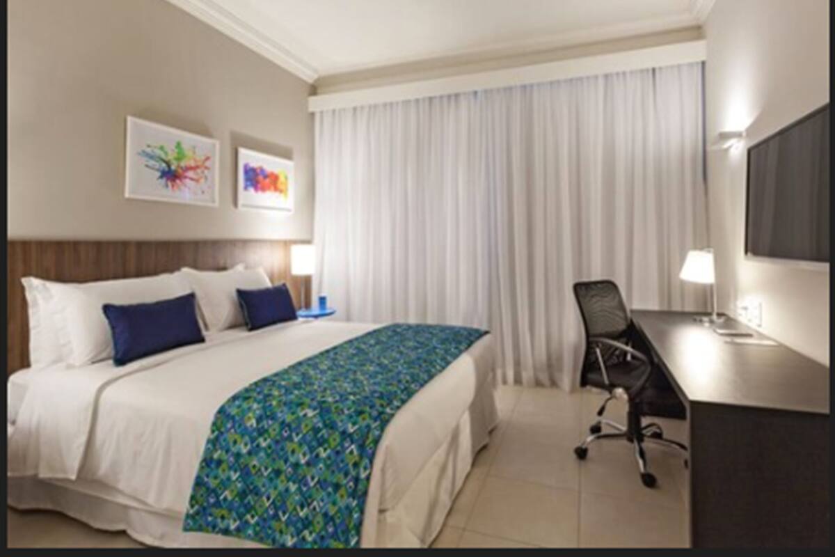 A hotel room with a bed, a desk, a chair, a TV, and two artwork pieces on the wall.