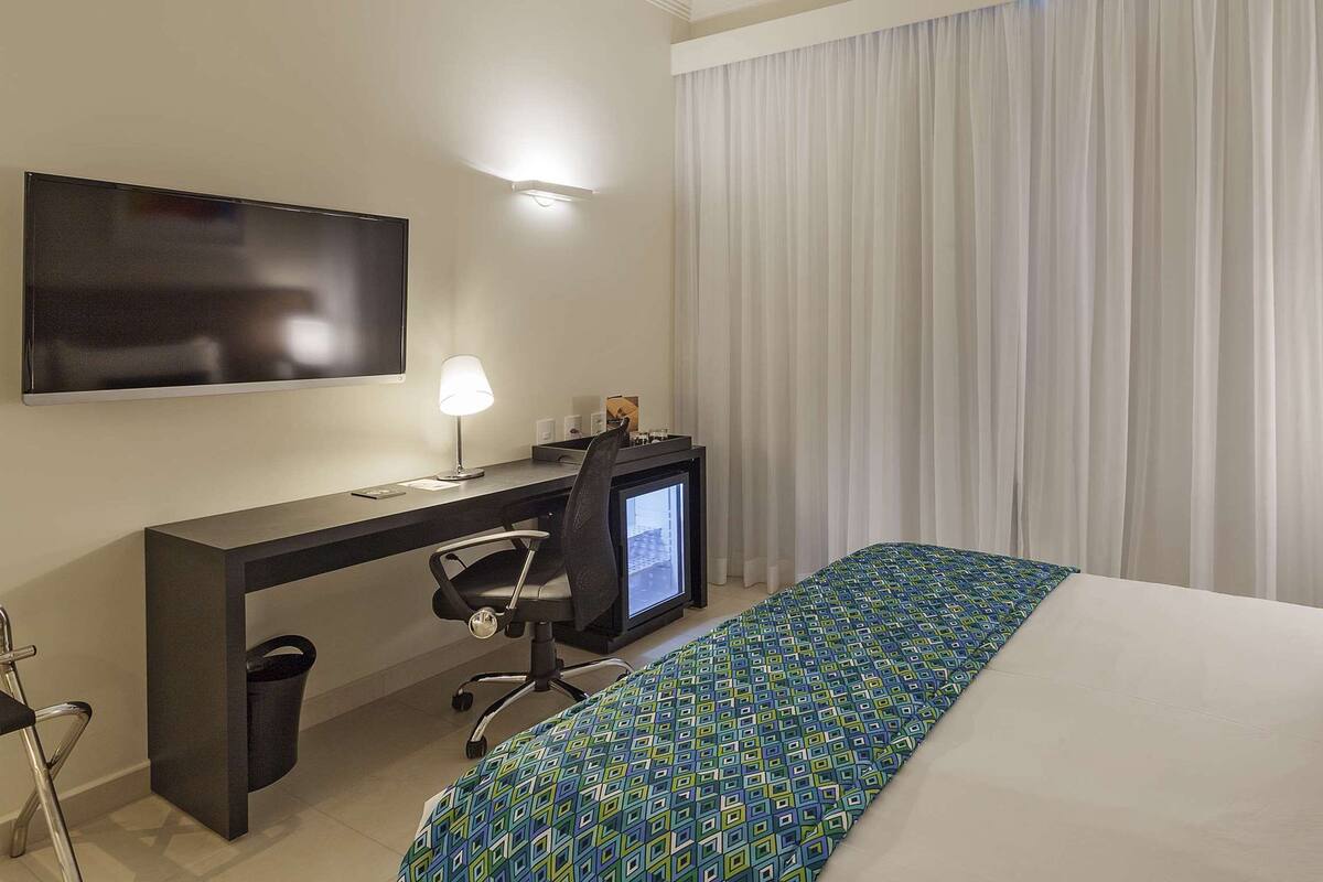 A hotel room with a bed, a desk with a computer, a chair, and a TV mounted on the wall.
