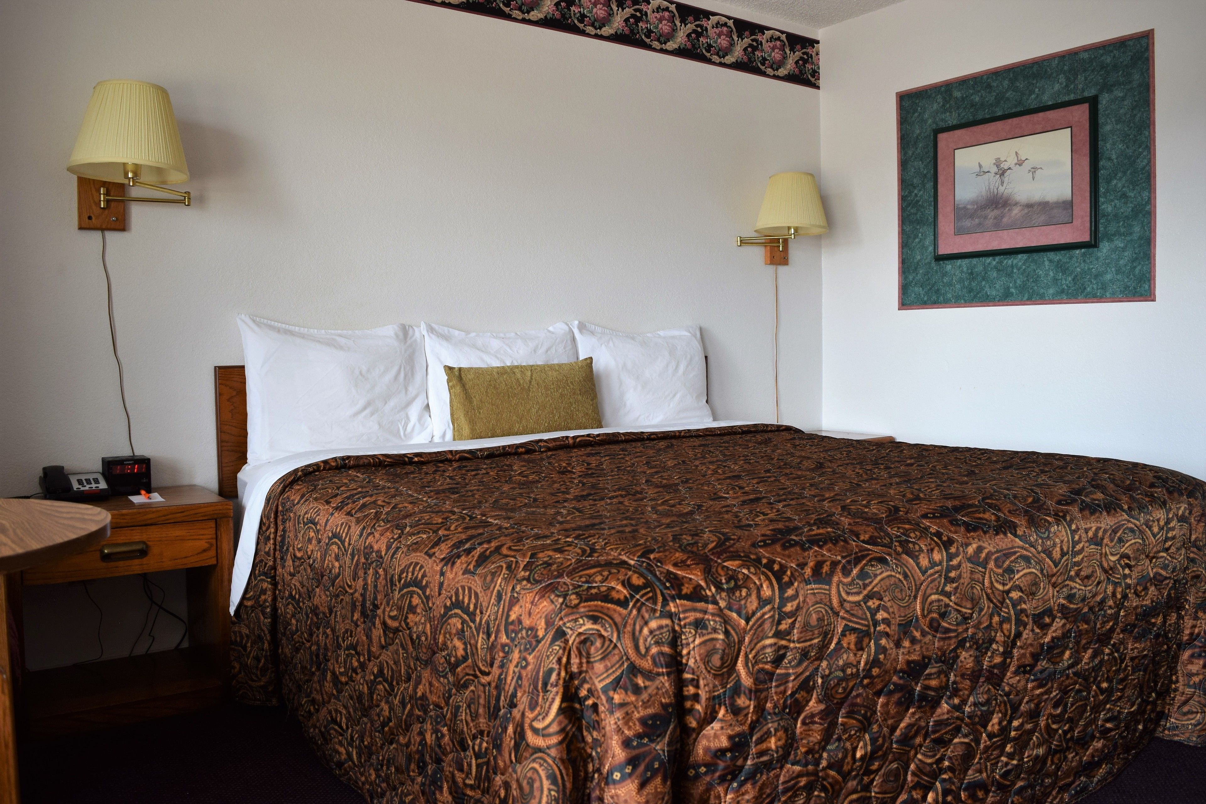 A neatly made bed with a patterned bedspread, two bedside lamps, a framed picture on the wall, and a small table with a radio.