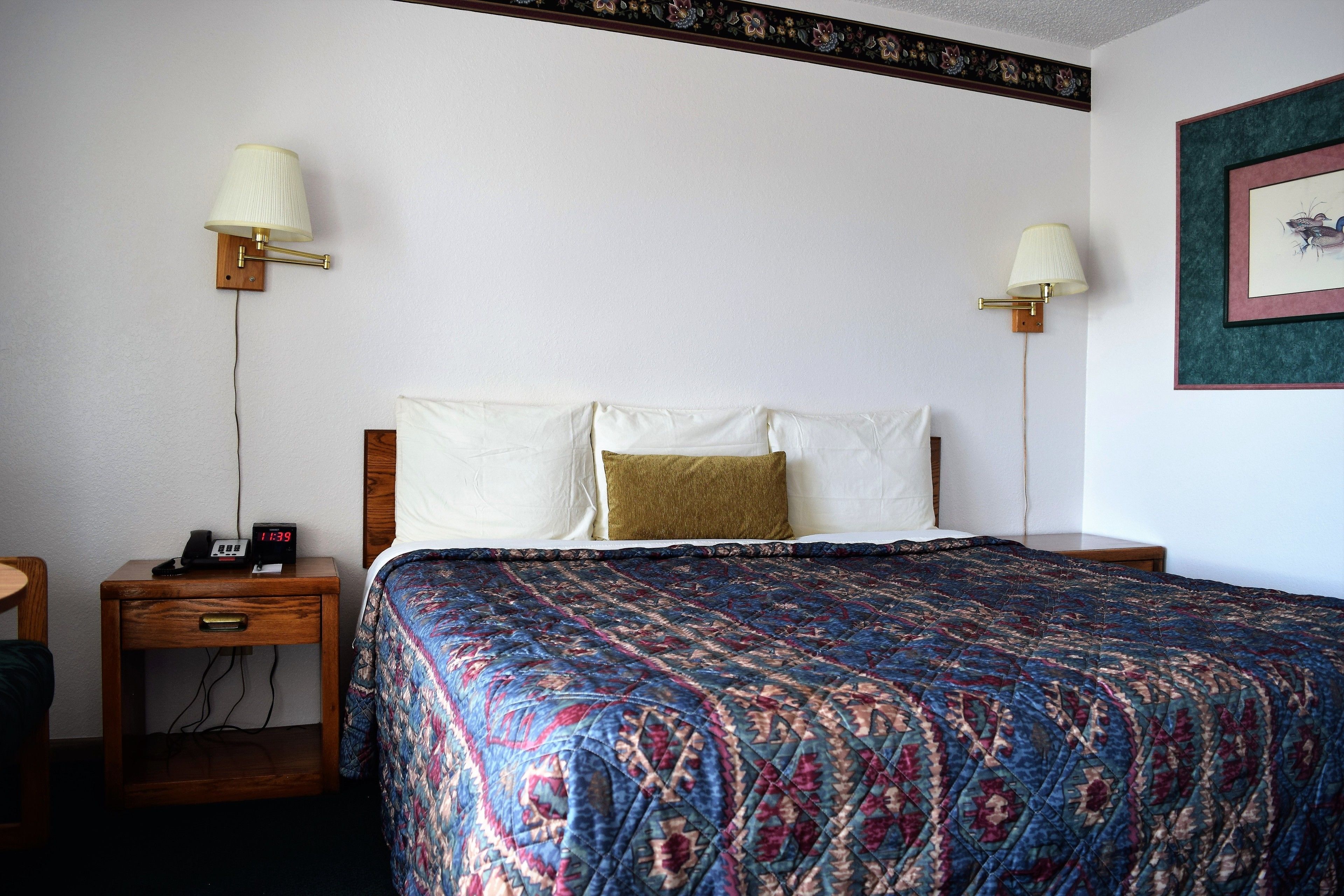 Habitación de hotel con cama, dos lámparas de noche, una mesita de noche con un reloj y un cuadro en la pared.